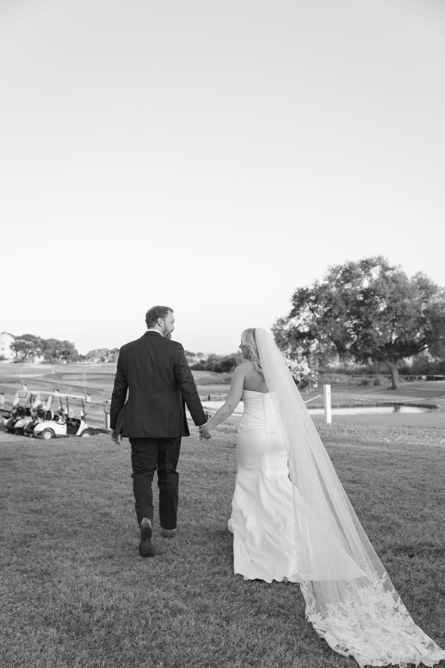 Black and white photo of bride and groom holding hands walking on golf course, bride wearing veil and wedding gown.