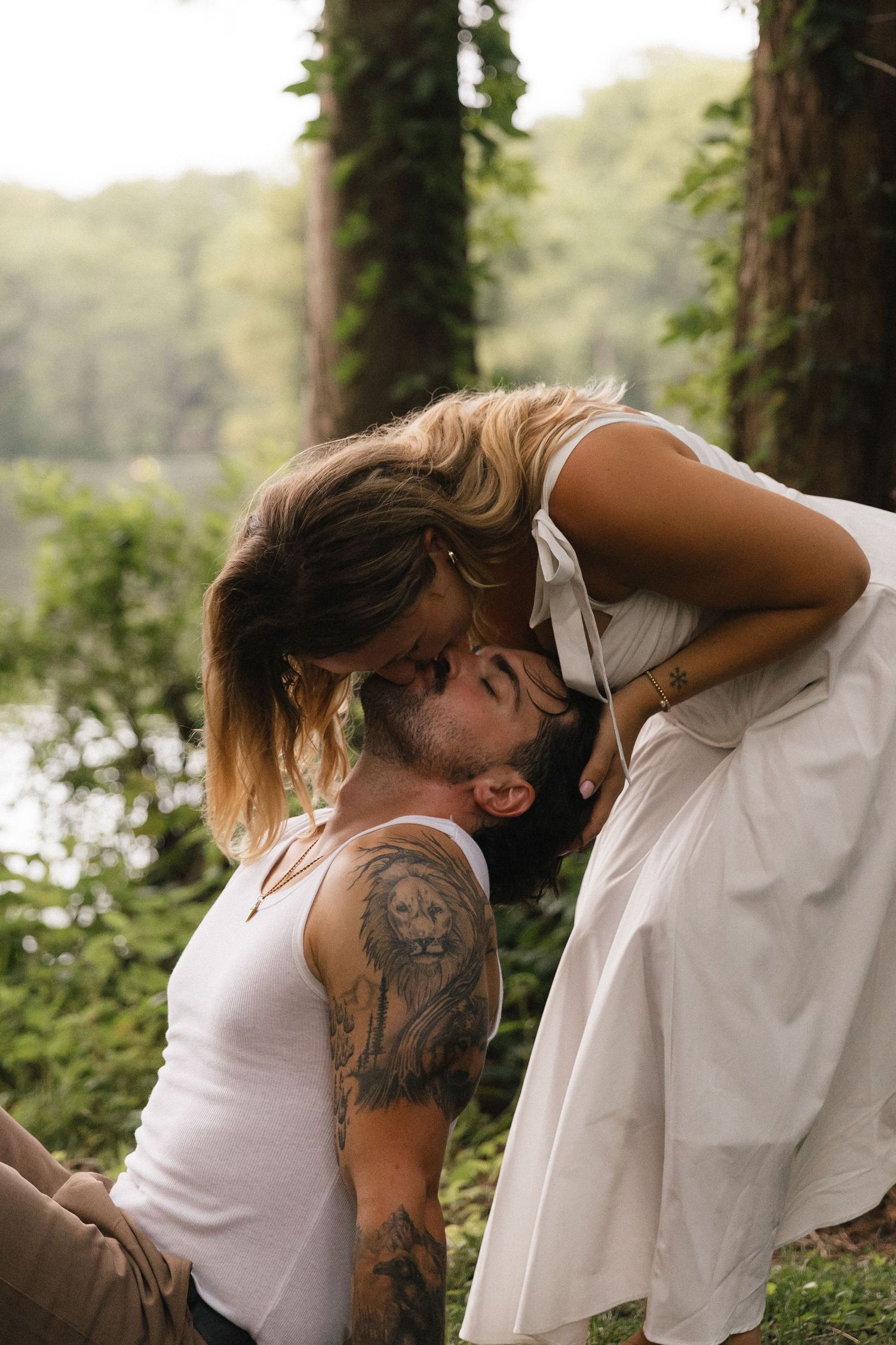 A couple shares a romantic kiss outdoors, man with lion tattoo in white tank, woman in white dress, green trees behind.