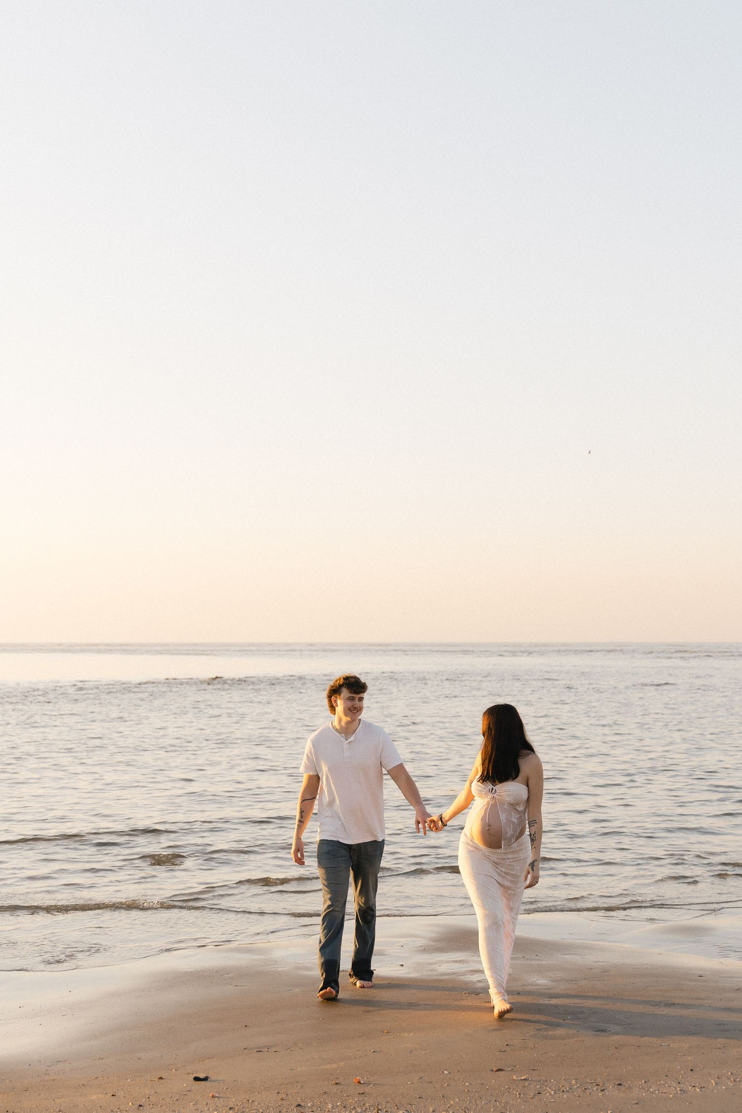 Couple holding hands walking barefoot on a sandy beach at sunset, wearing casual white outfits.