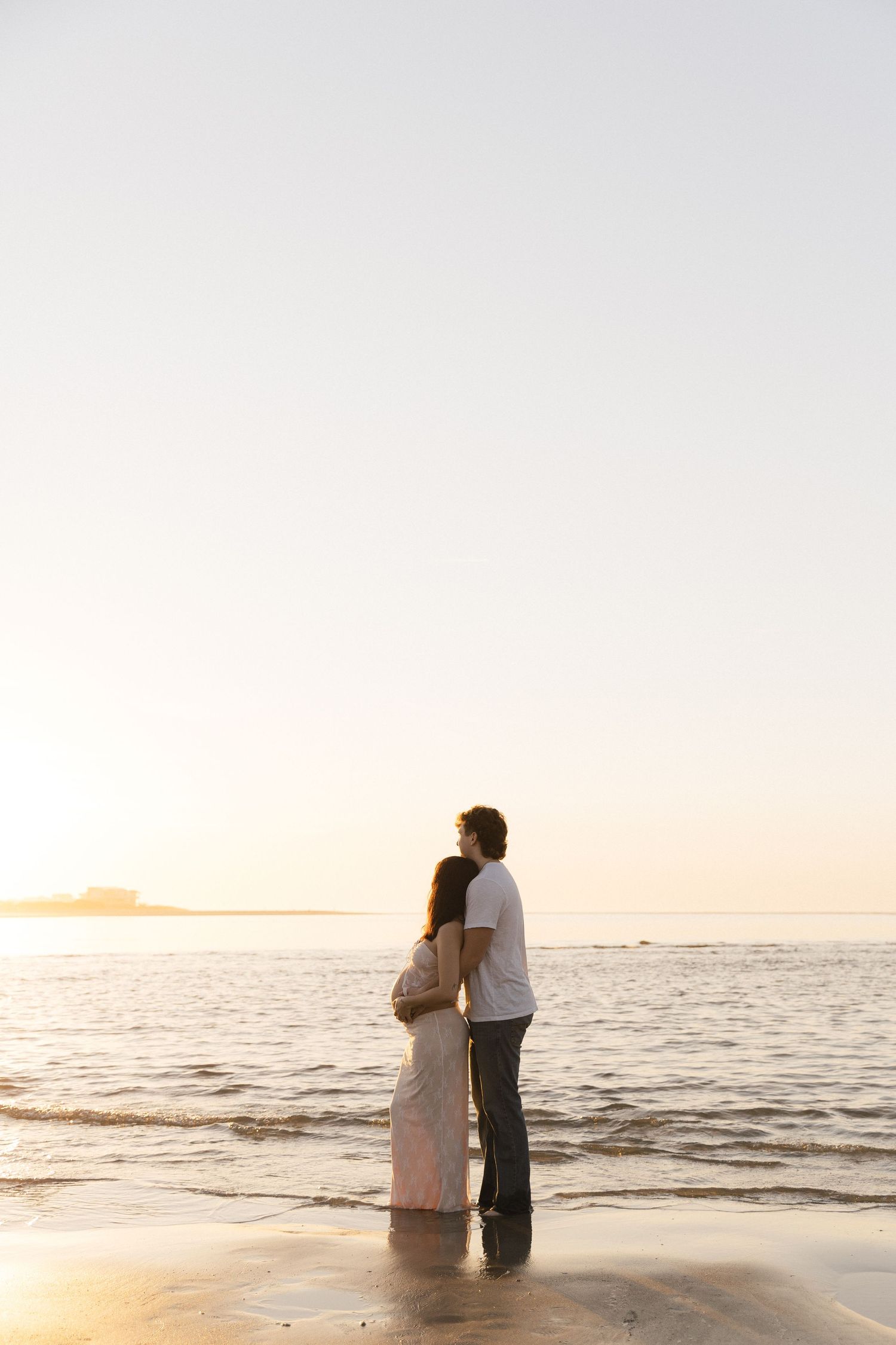 Couple embracing on a sandy beach at sunset, standing at the water's edge with a calm ocean horizon.