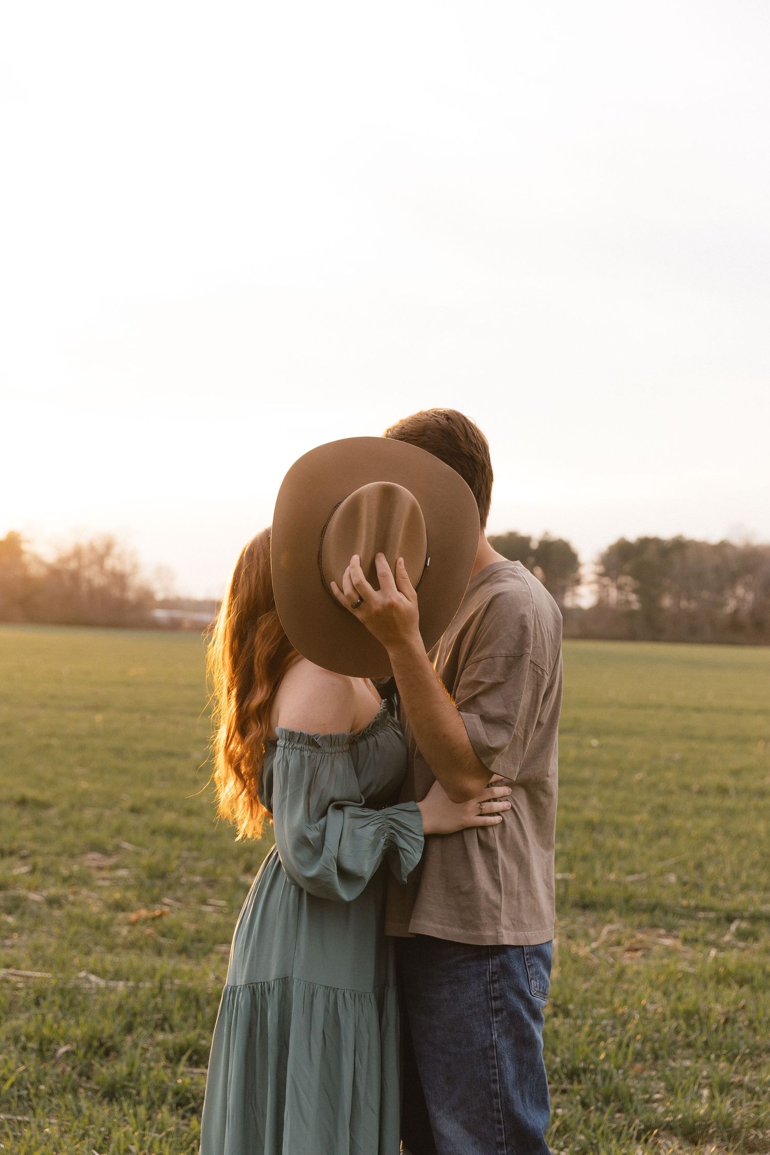 Couple kissing behind a brown hat in a green field at golden hour, woman in teal dress, man in gray shirt.