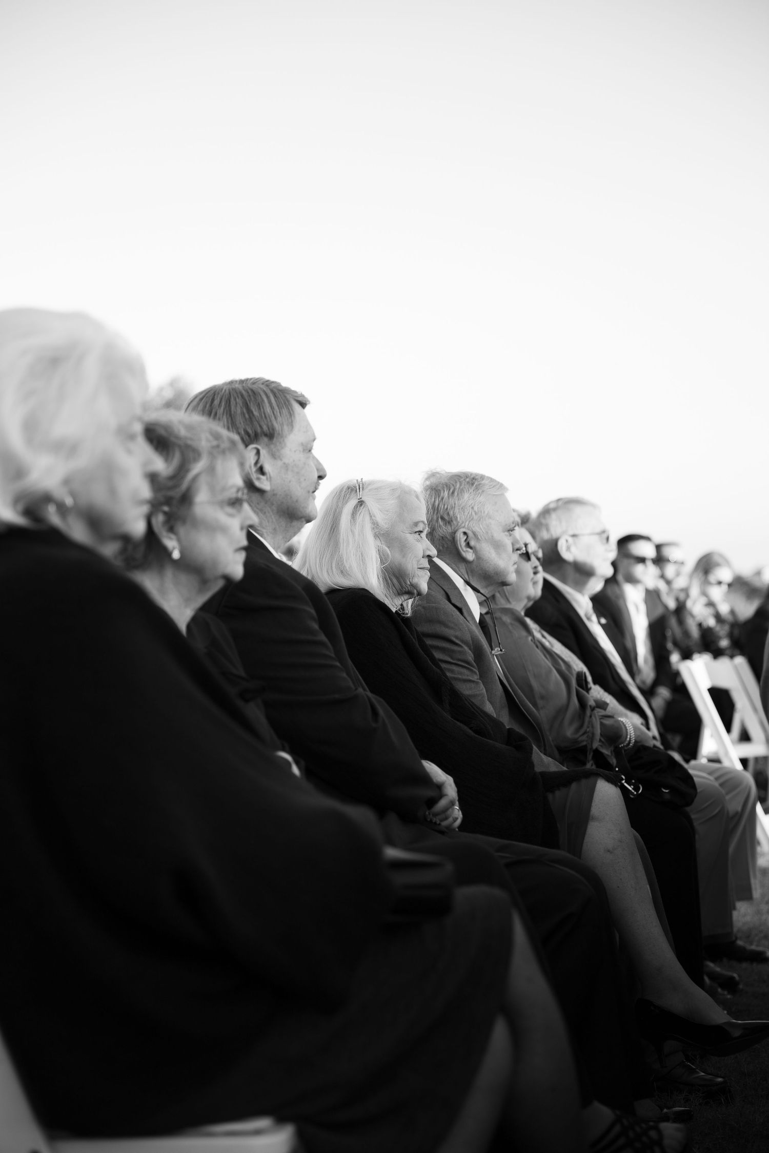 Black and white photo of elegantly dressed guests seated in rows at a formal outdoor ceremony.