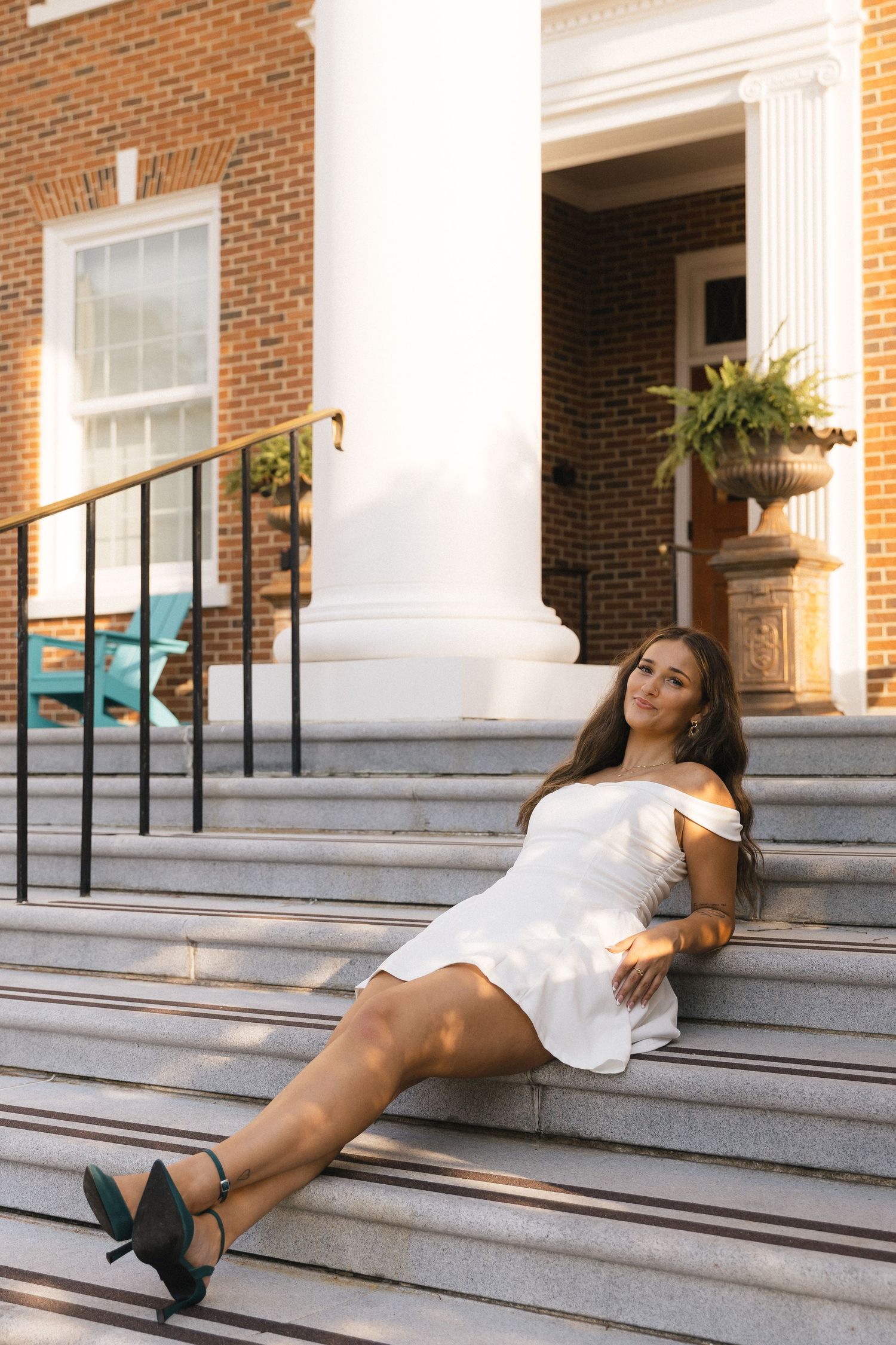 Young woman in white off-shoulder dress poses gracefully on stone steps of a classic brick building in golden hour light.
