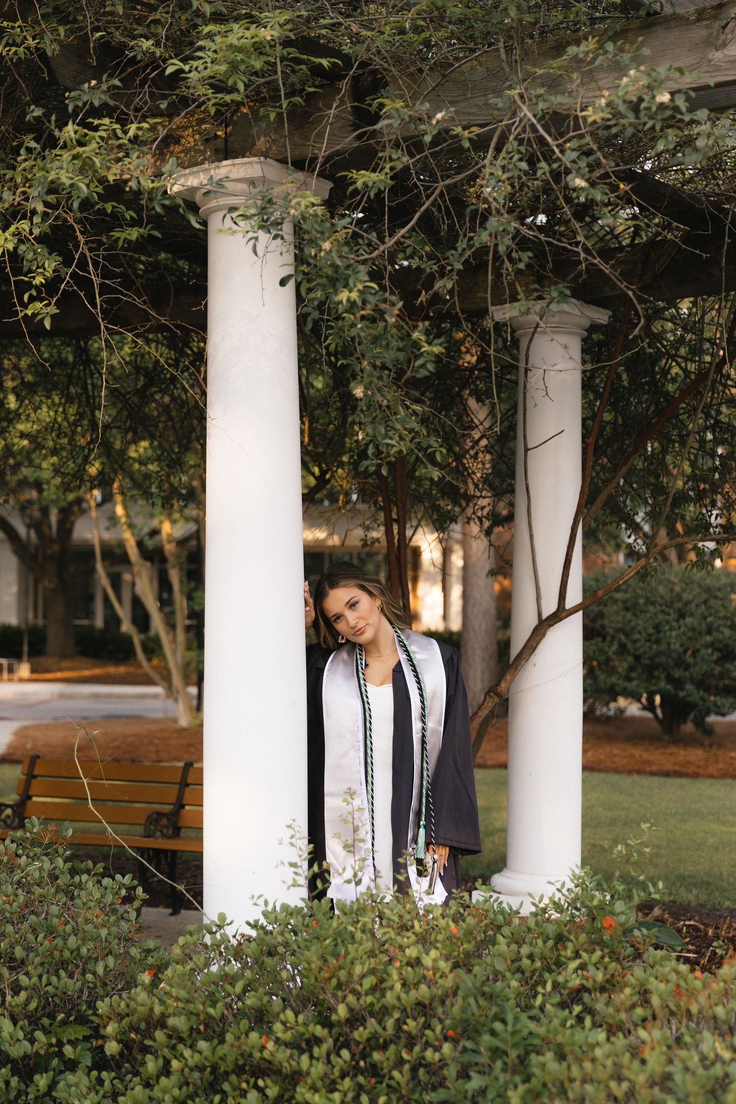 Graduate in cap and gown poses between white columns surrounded by lush green trees in an outdoor campus setting.