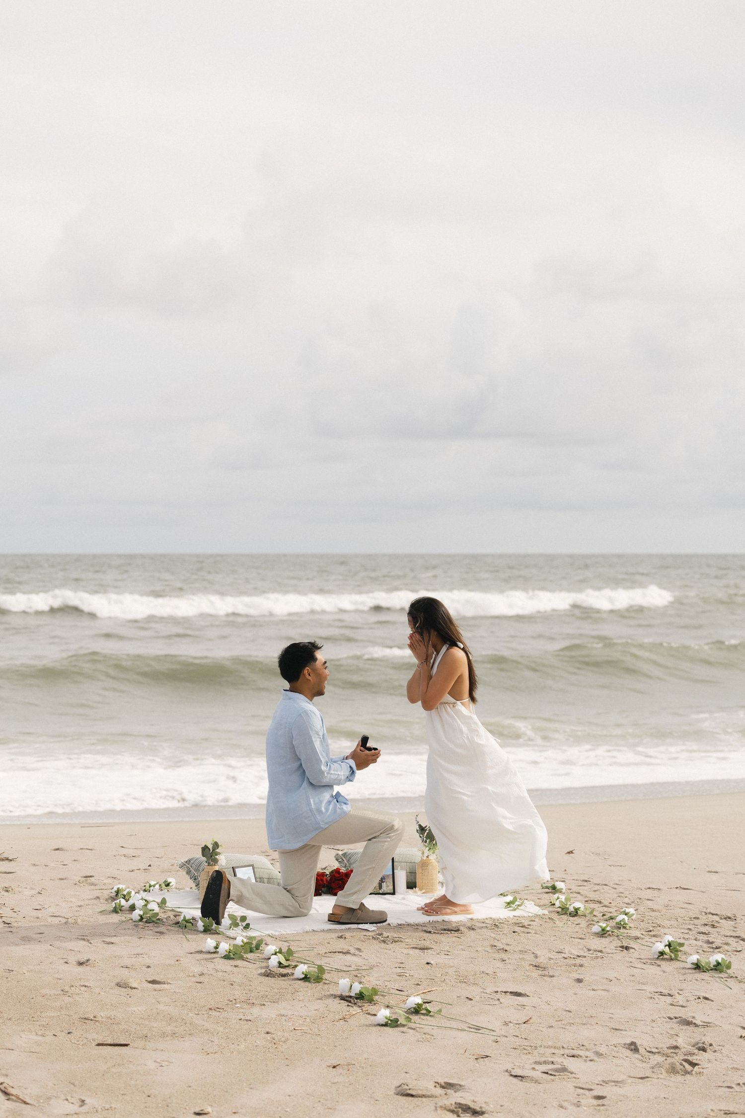 Man proposing to woman on a beach with flower decorations, ocean waves in the background.