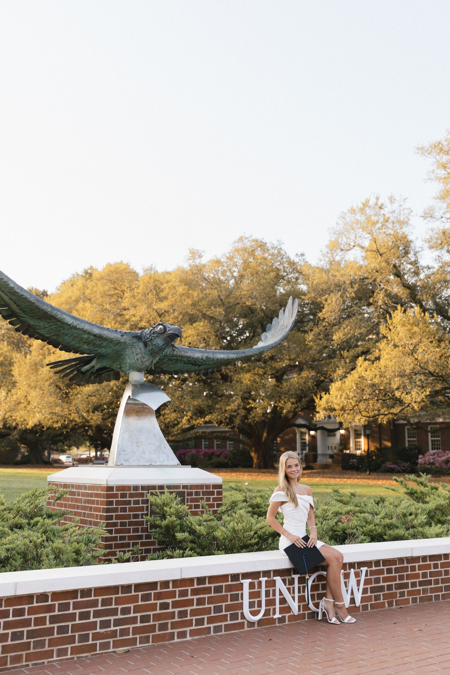 A young woman sits on a brick wall beside the UNCW Seahawk statue surrounded by golden trees.