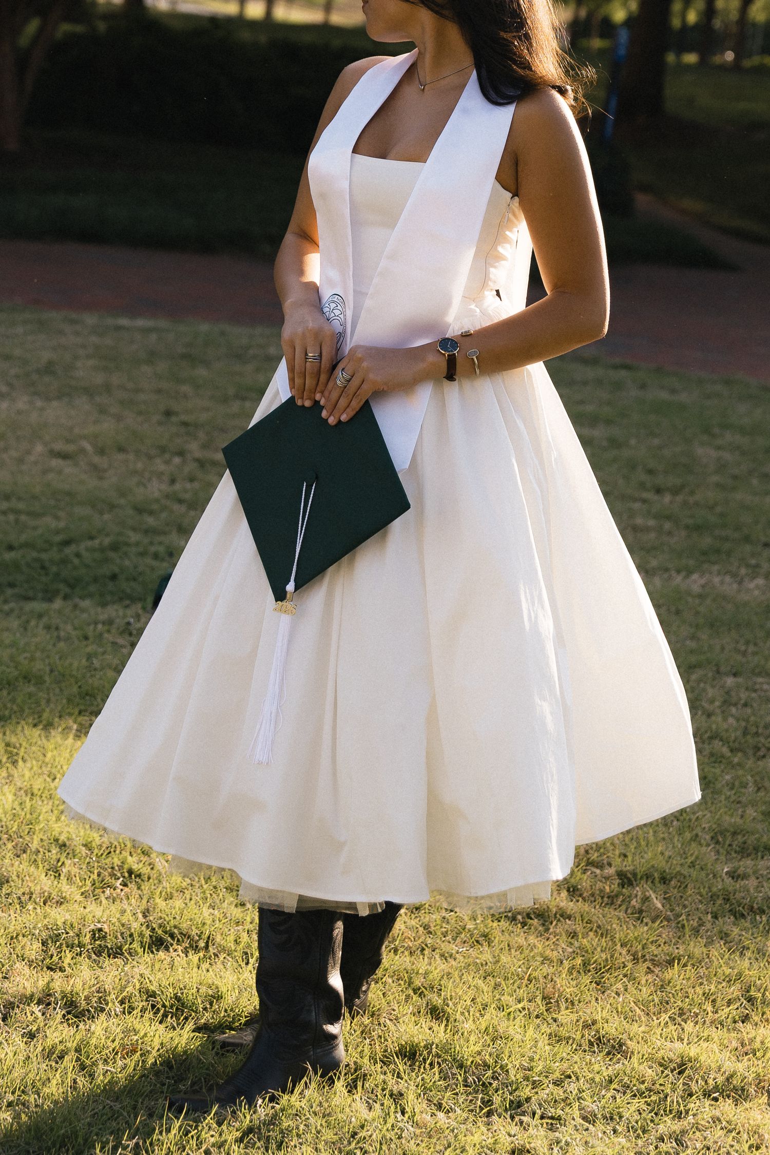 Young woman in white midi dress holding graduation cap, standing on green grass in outdoor park setting.
