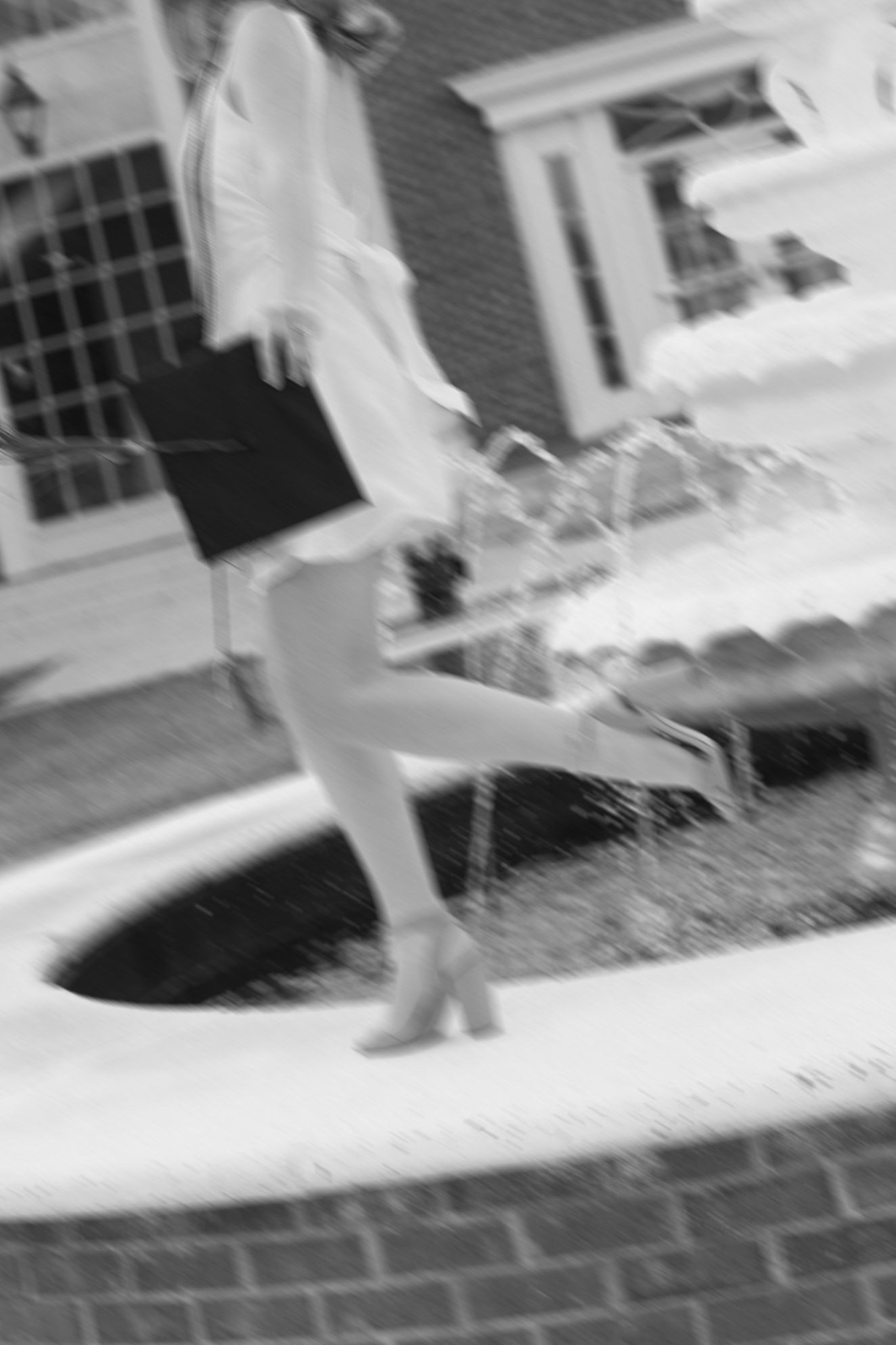 Blurred black and white photo of a woman in heels standing by an outdoor fountain near a brick building.