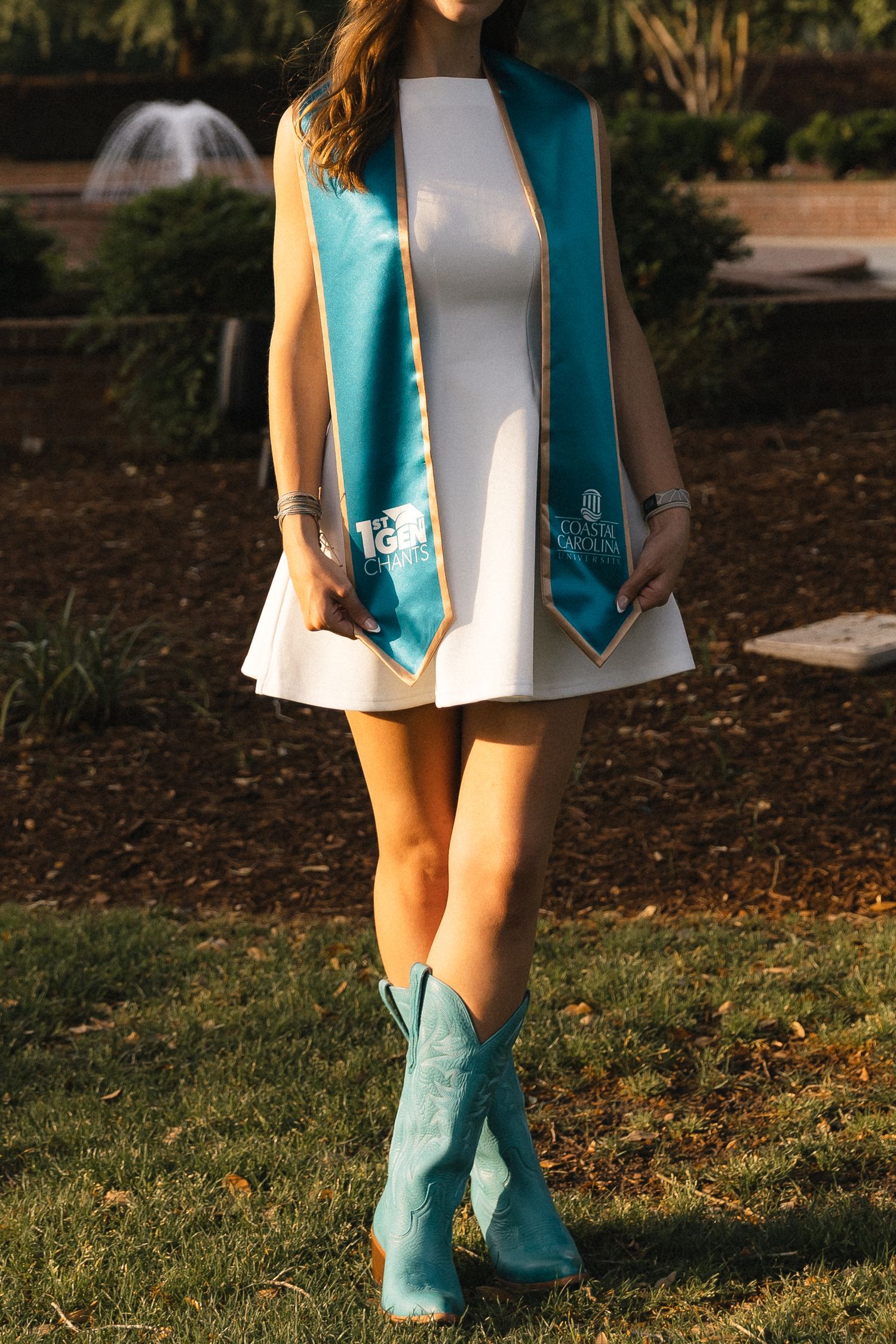 Graduate in white dress with teal sash and matching cowboy boots posing outdoors on green grass.