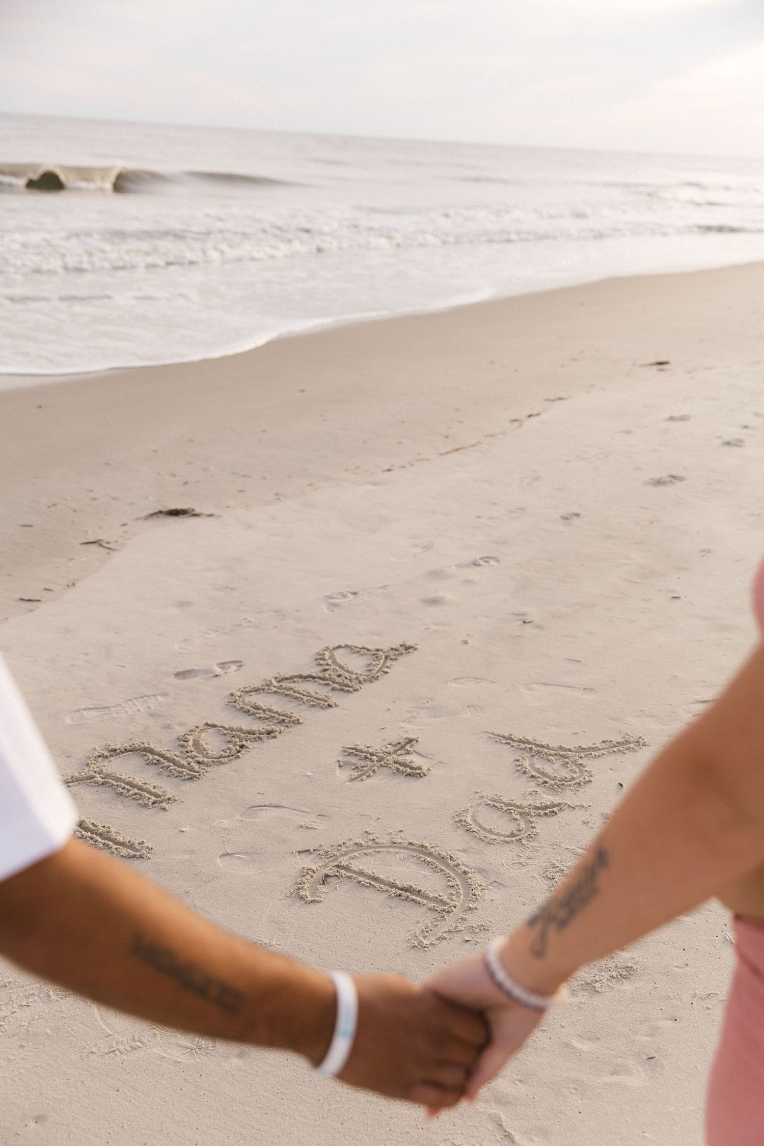 Couple holding hands on beach with 'Mama + Dad' written in sand near ocean waves.