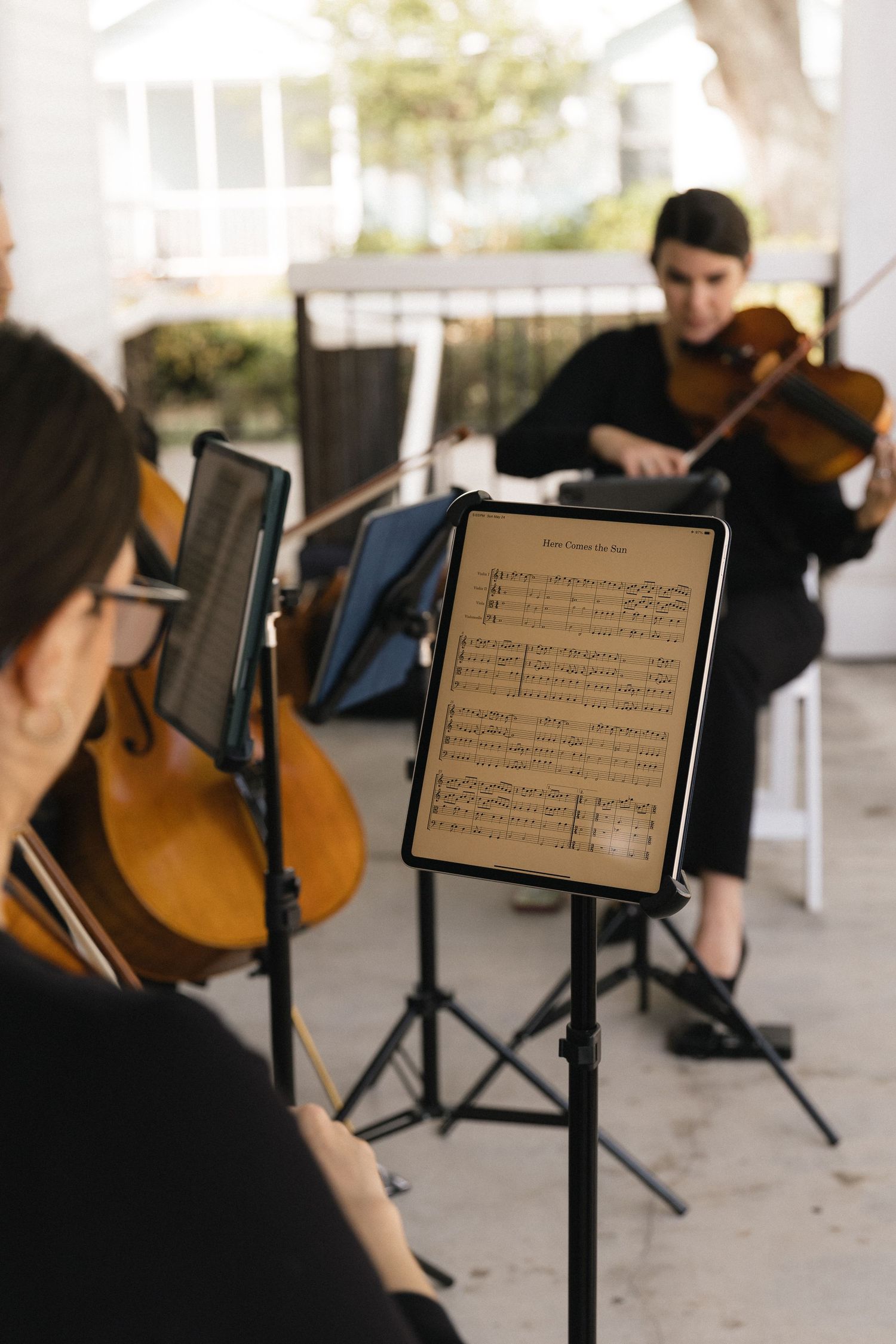 Two musicians in black attire play string instruments outdoors, with sheet music displayed on a tablet stand.
