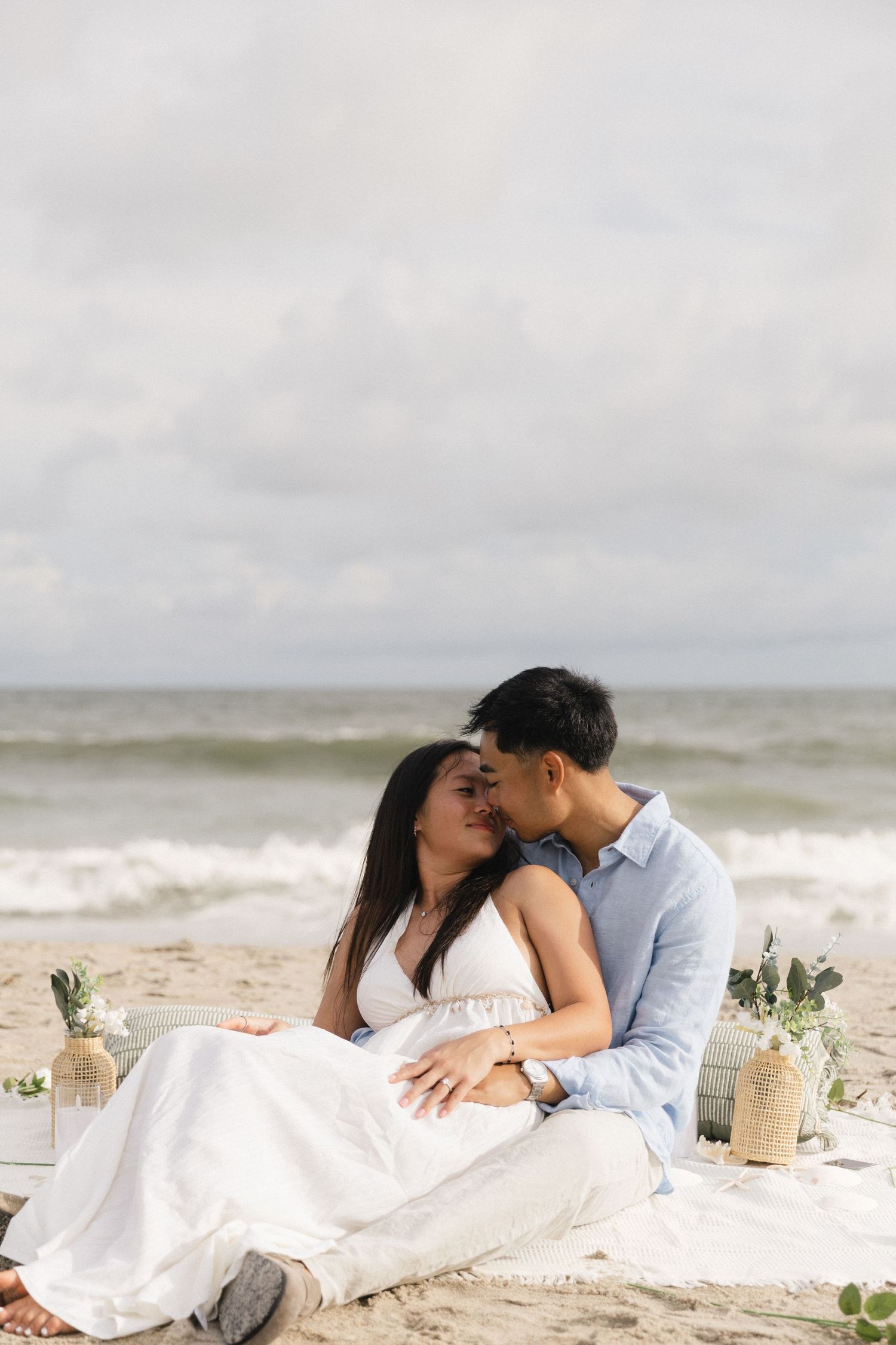 Couple sharing a romantic kiss on a sandy beach with ocean waves and cloudy sky backdrop.