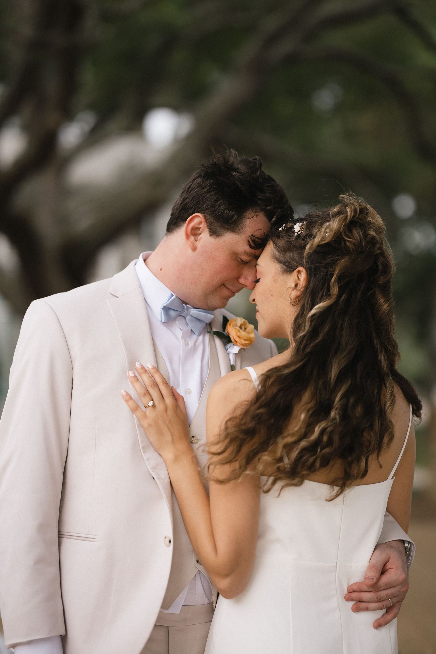 Bride and groom share a romantic forehead touch in outdoor wedding portrait, groom in cream suit with blue bow tie.