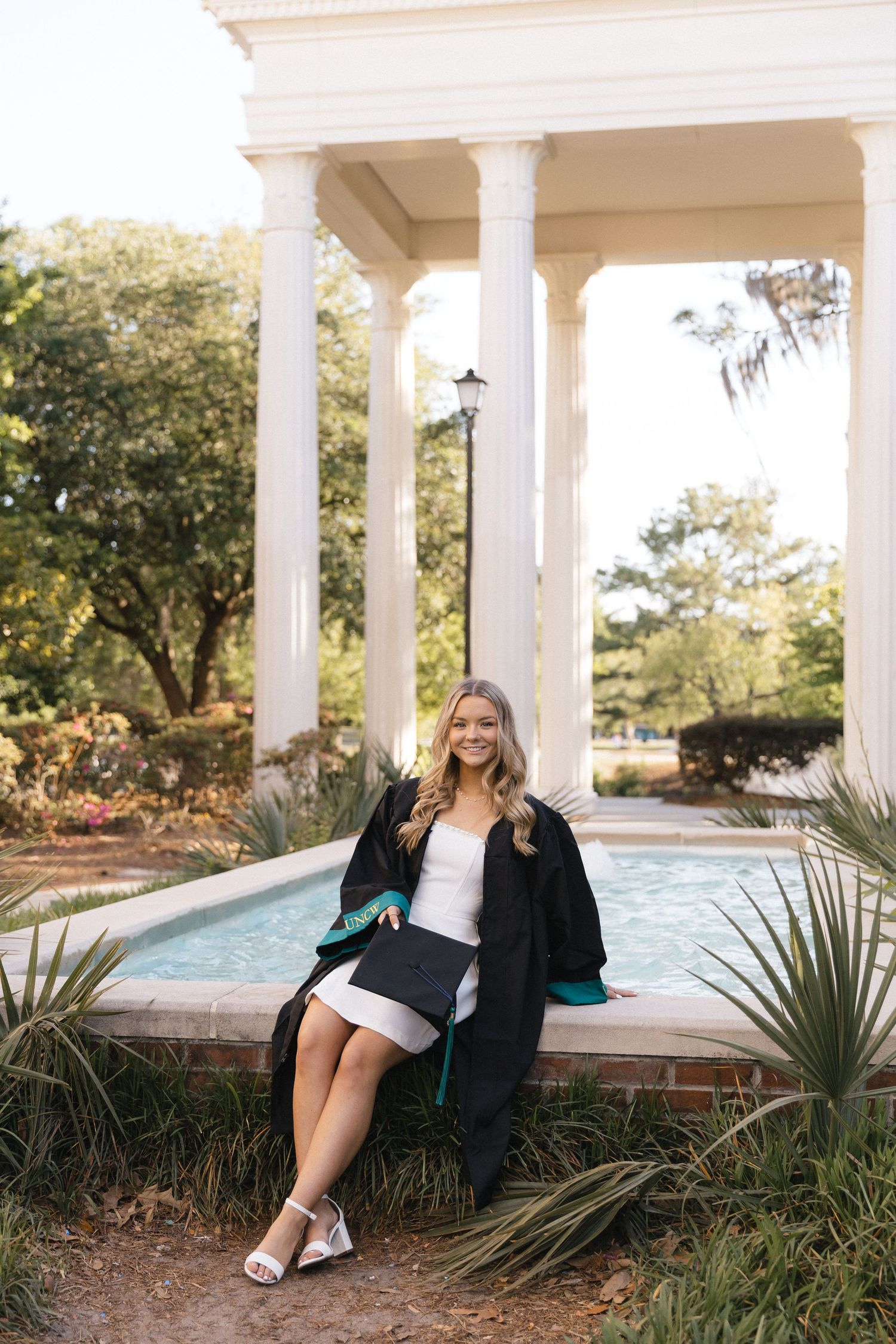 A smiling graduate in black gown sits by a fountain with white columns and lush greenery in the background.