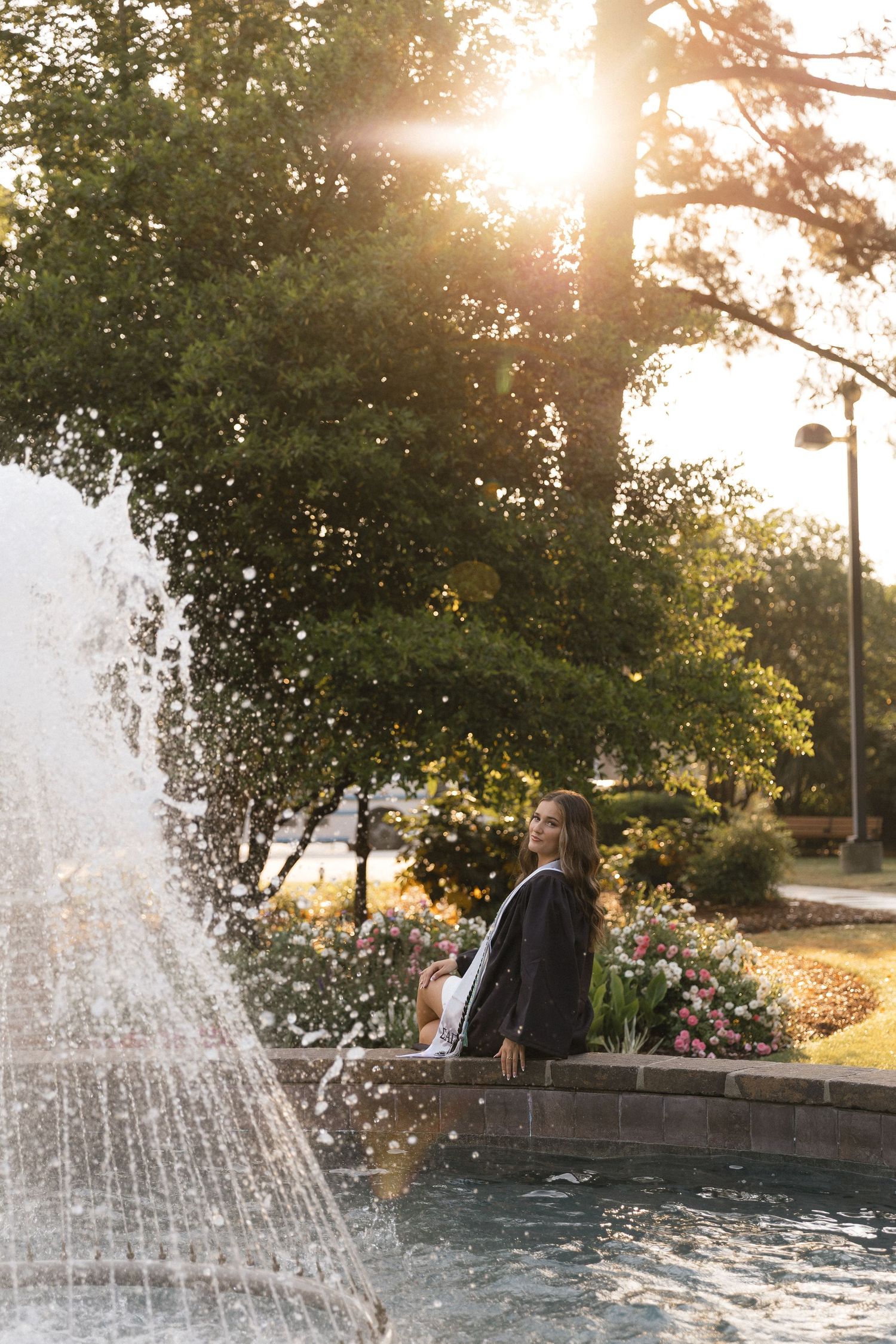 A graduate in cap and gown sits by a sunlit park fountain surrounded by lush green trees and colorful flowers at golden hour.
