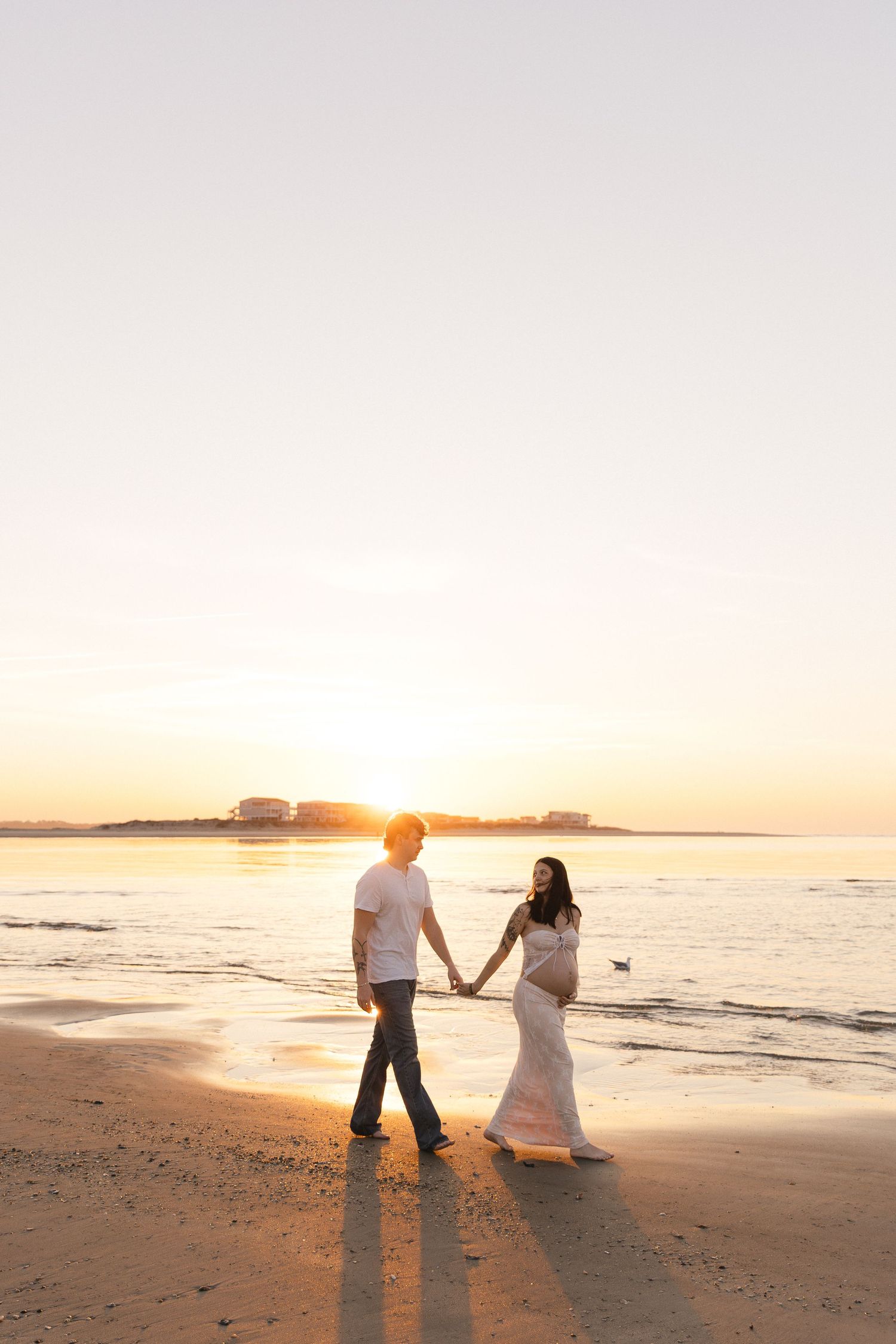 Pregnant couple holding hands on a sandy beach at golden sunset, woman in white dress cradling baby bump.