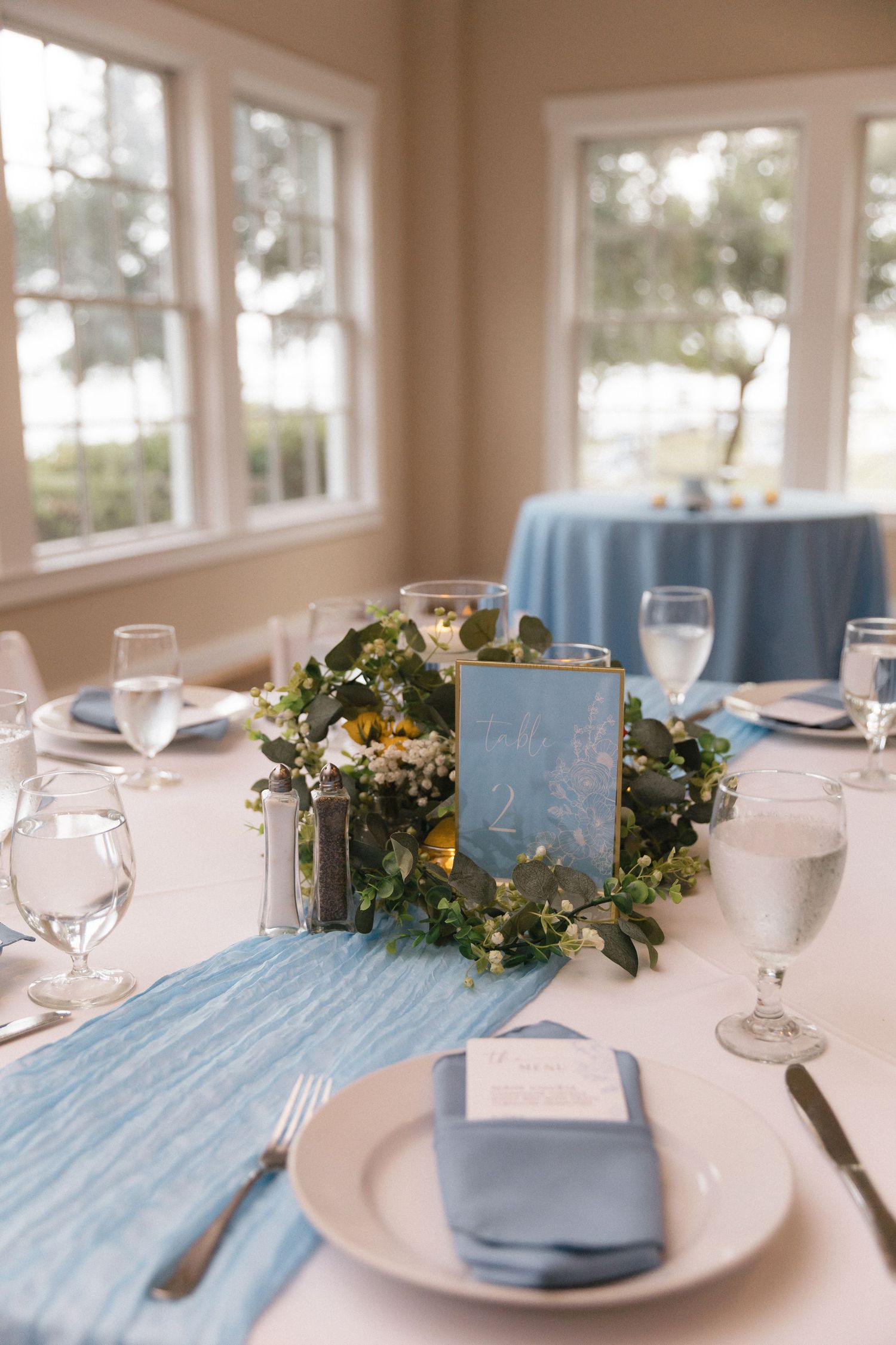 Elegant wedding reception table with blue runner, greenery centerpiece, candles, and place settings by sunlit windows.