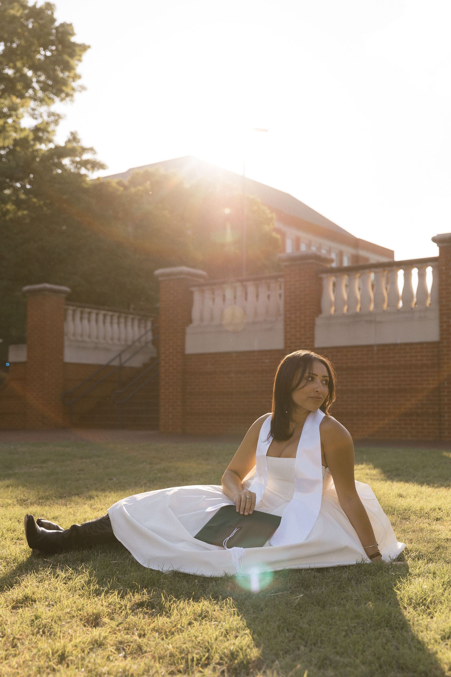 Young woman in white dress sitting on grass holding graduation cap with sunlit brick building behind her.