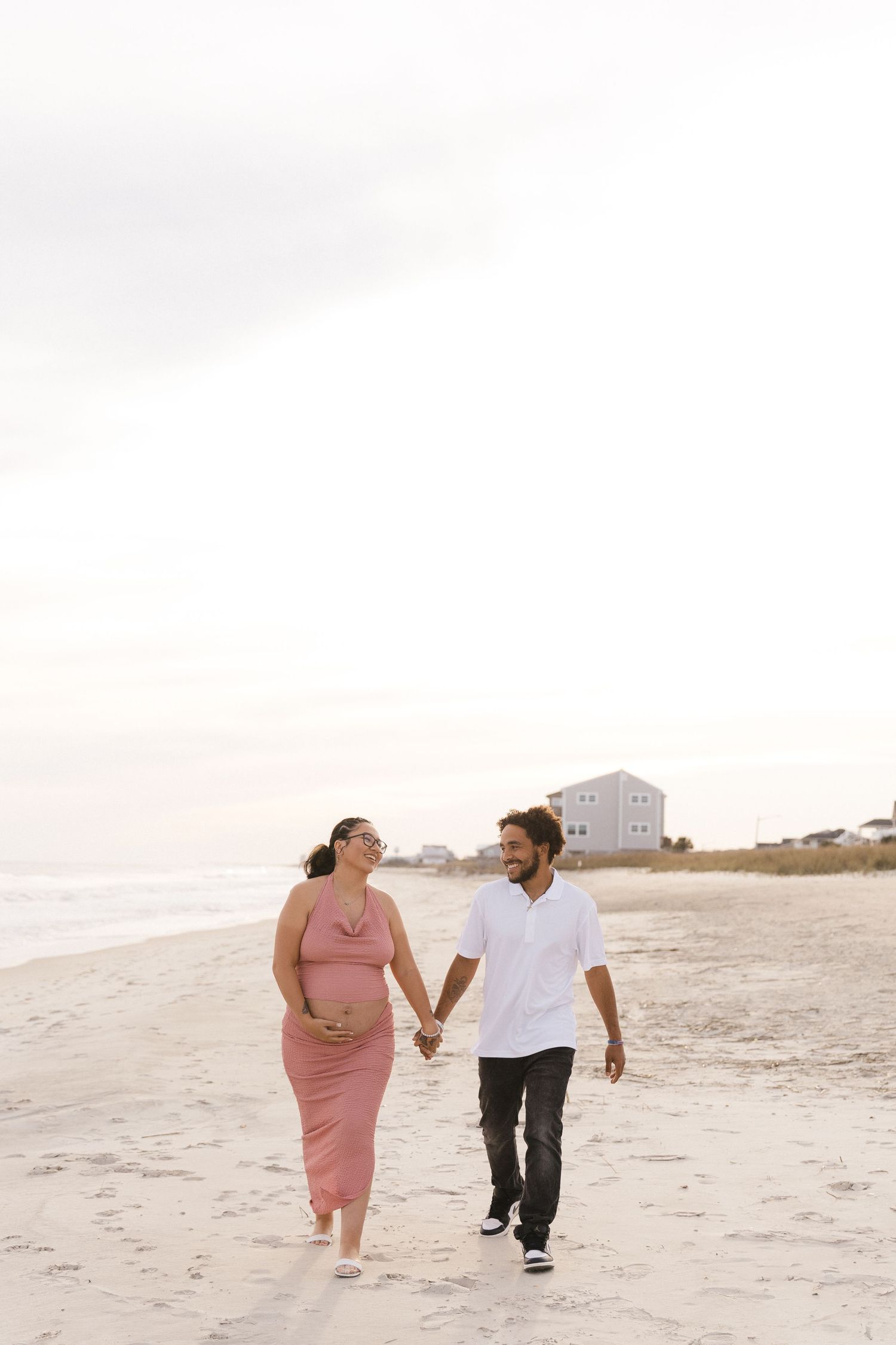 Pregnant couple holding hands walking on sandy beach, woman in pink dress, man in white shirt, ocean backdrop.