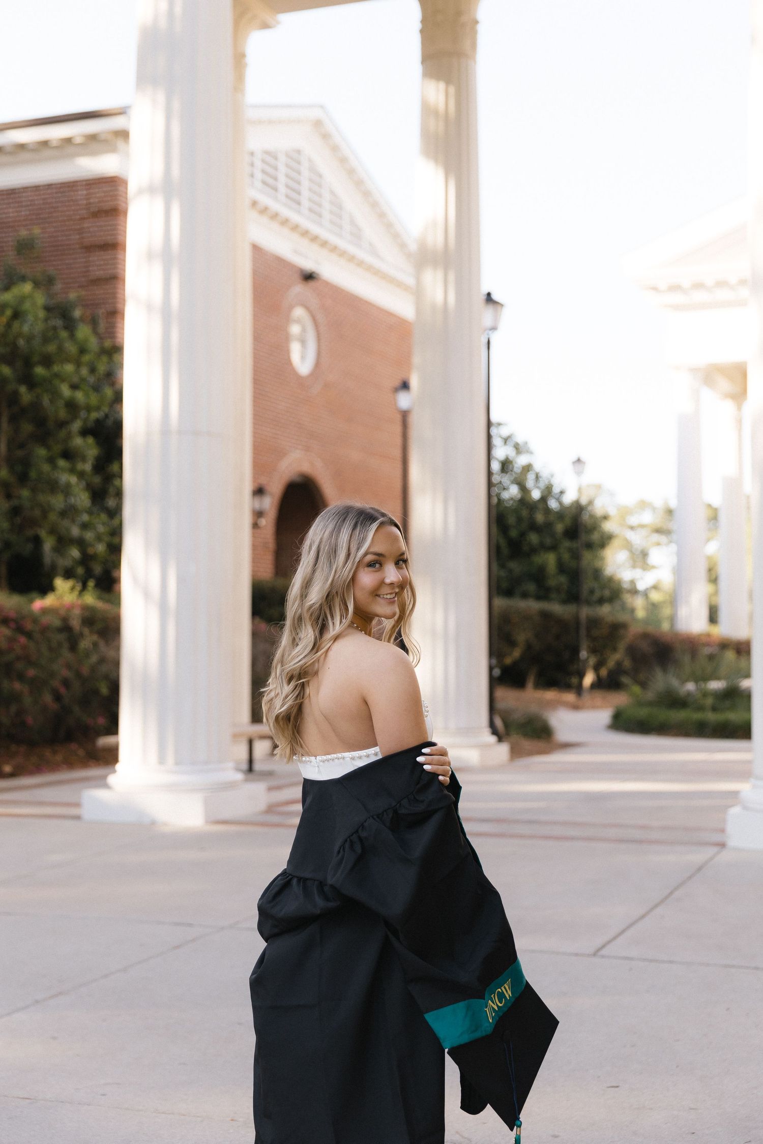 Smiling blonde woman in graduation gown holding cap, posing in front of brick building with white columns.