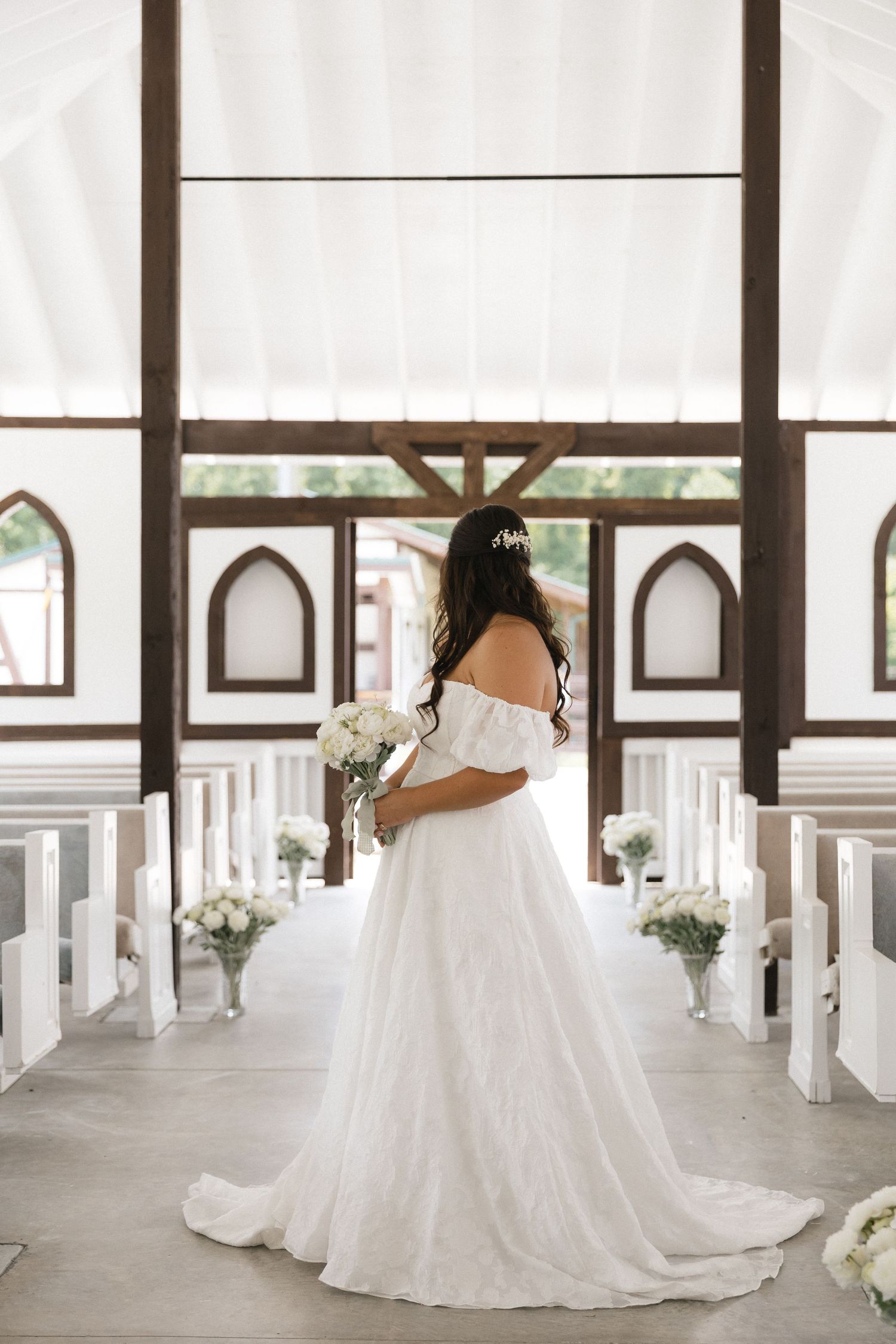 Bride in off-shoulder white gown holds bouquet at chapel altar aisle with white pews and floral decor.