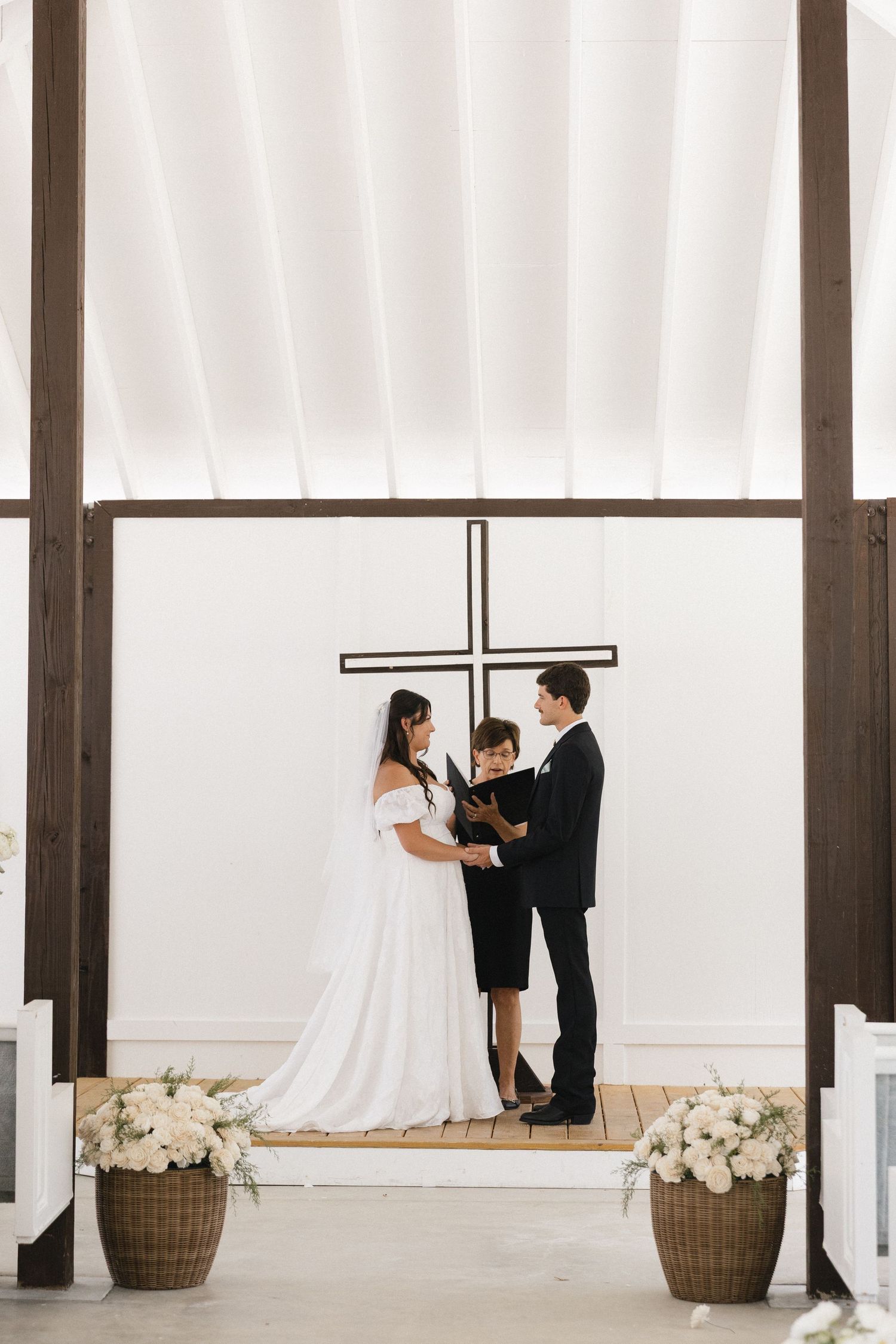 Bride and groom exchange vows before a wooden cross in a bright white chapel with floral arrangements.