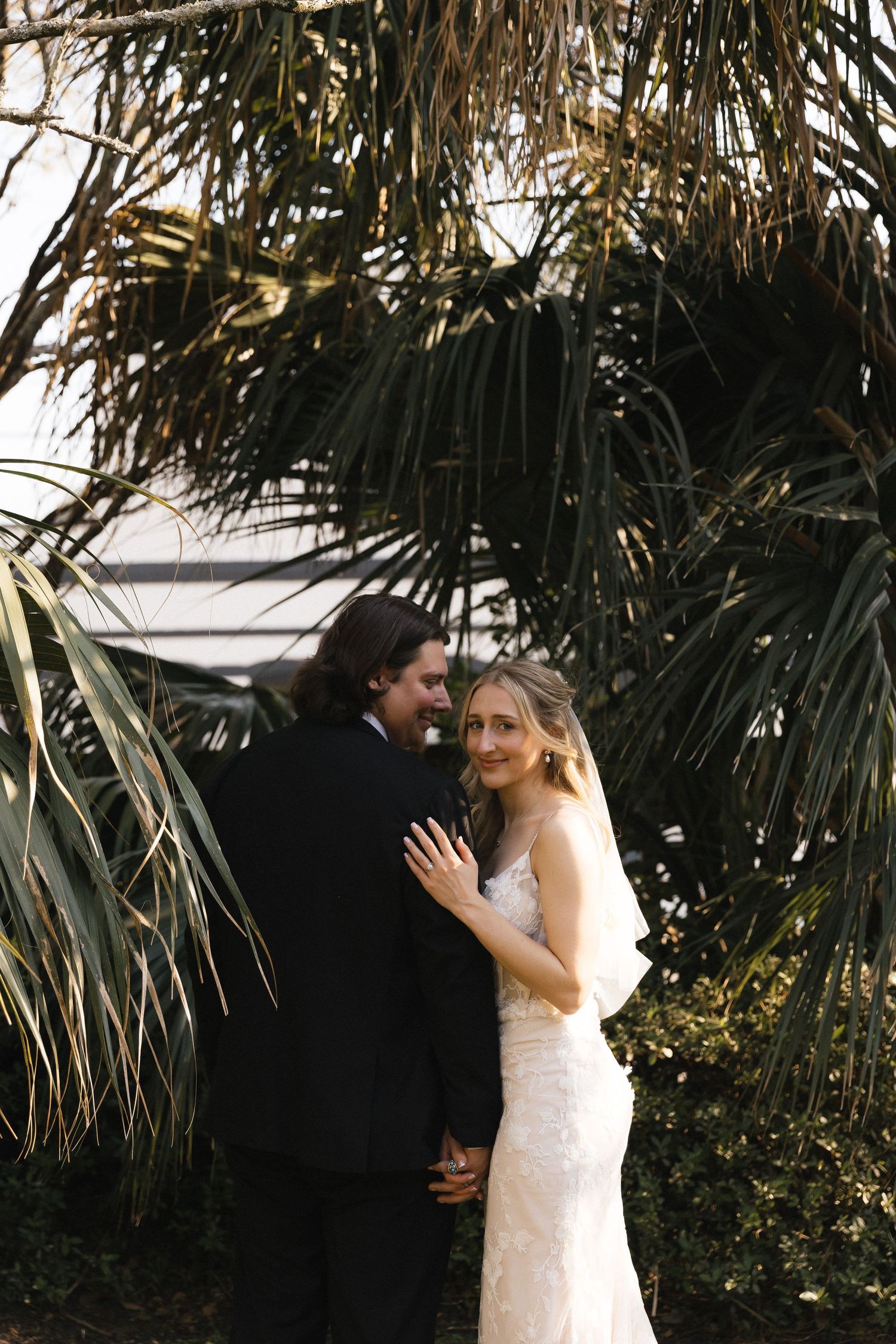 Bride in lace gown embraces groom in black tuxedo among tropical palm trees in outdoor wedding portrait.