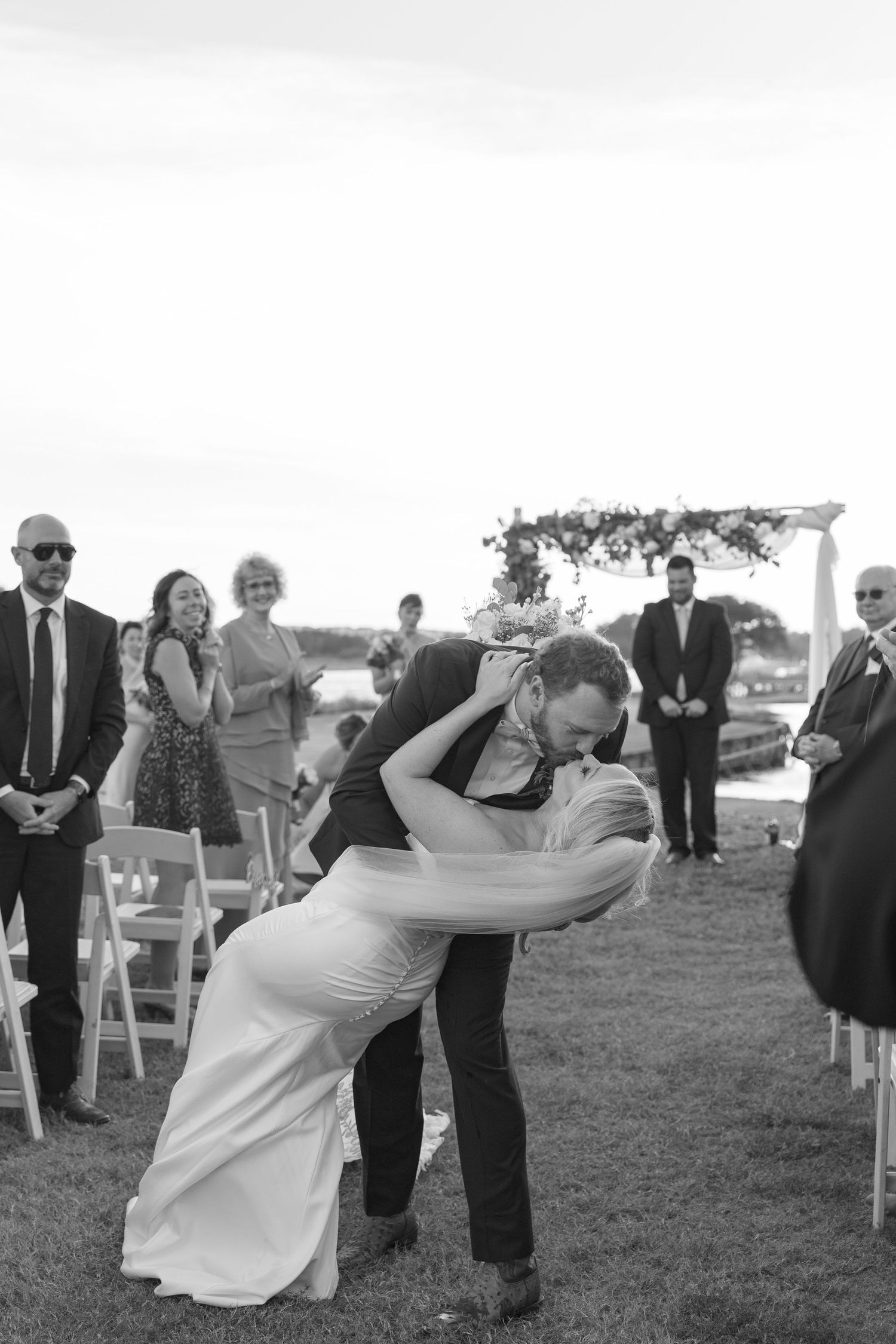 Black and white photo of groom dipping bride for a kiss after outdoor wedding ceremony with guests applauding.