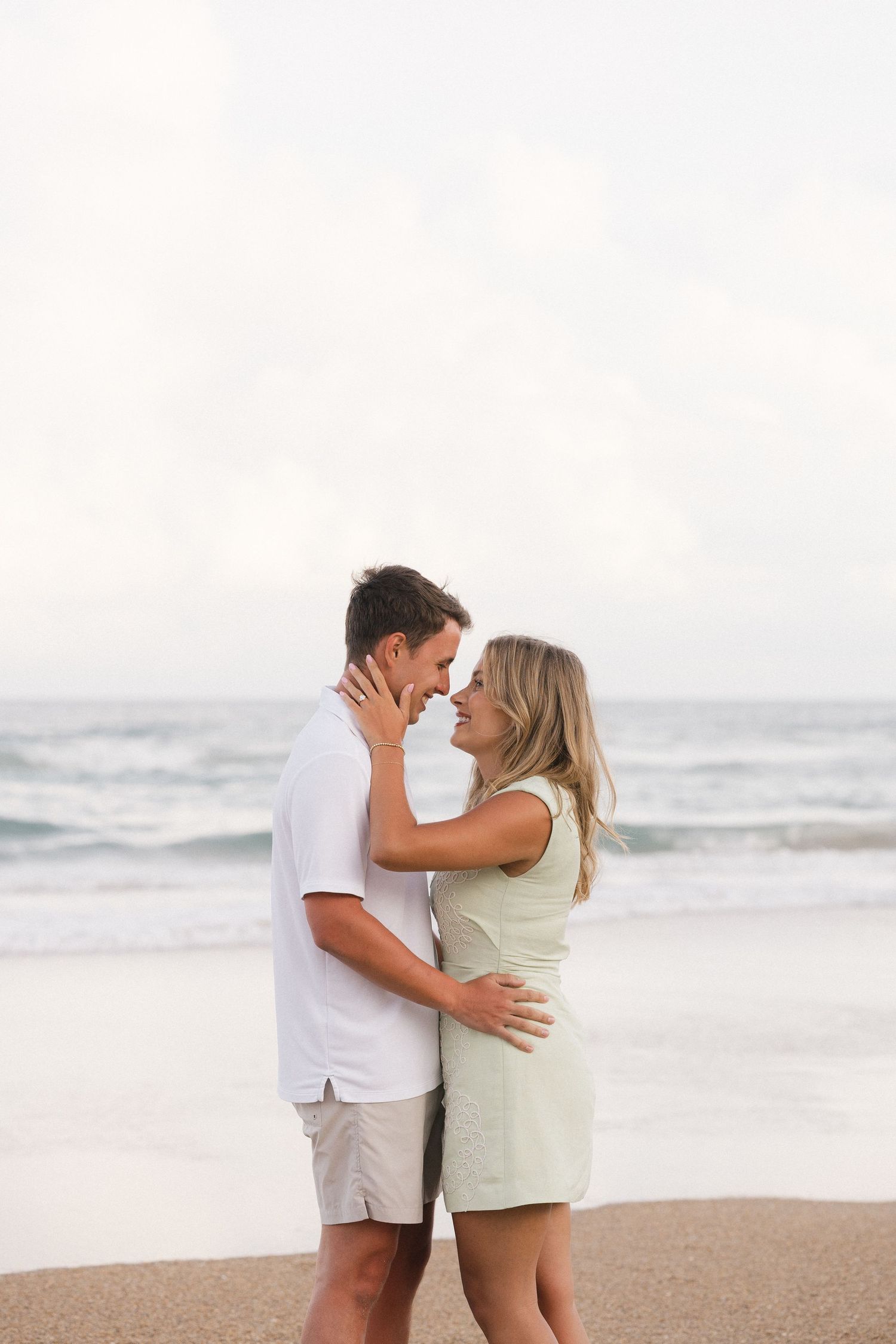 Romantic couple embracing and smiling at each other on a sandy beach with ocean waves in the background.