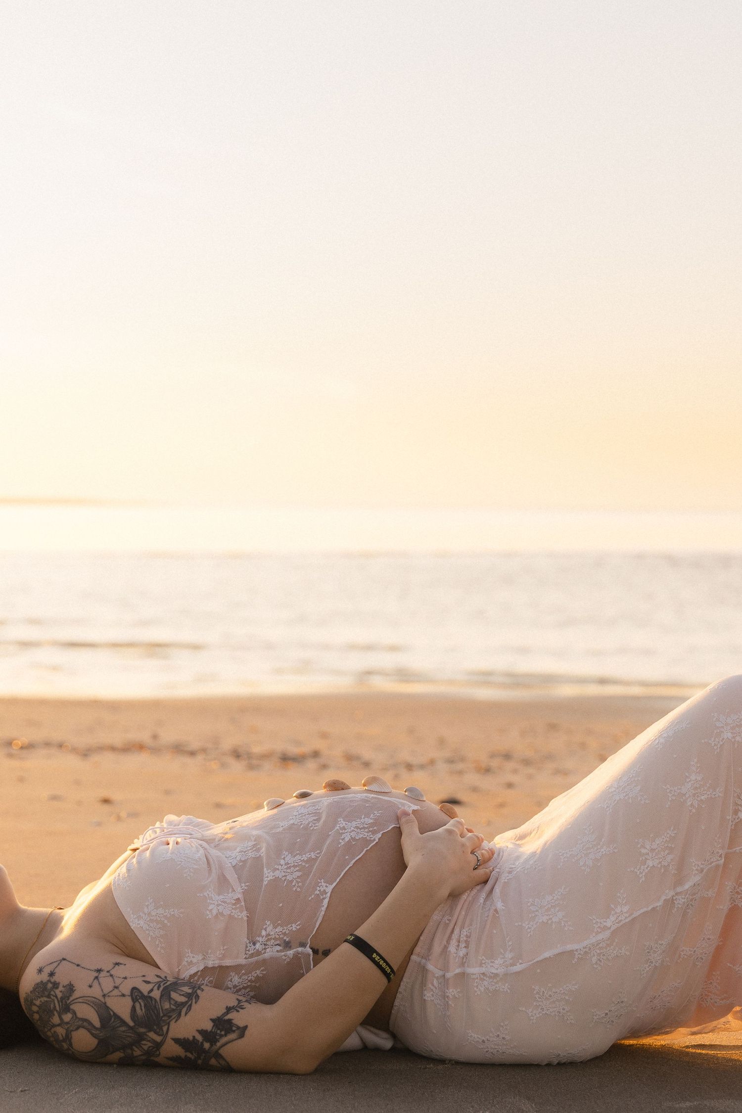 Pregnant woman in white lace dress lying on sandy beach at sunset, showing tattoo on arm.