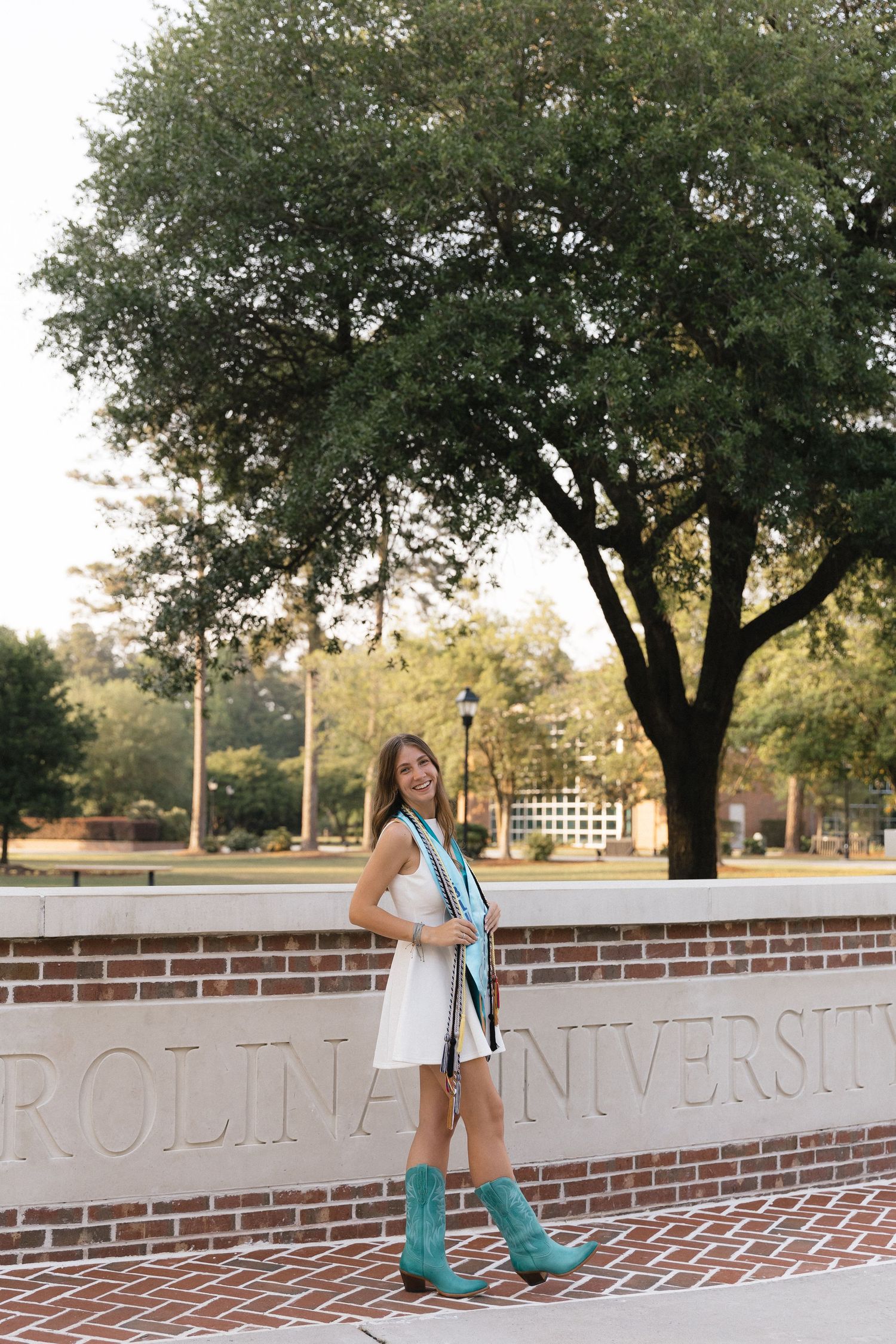 A smiling young woman in a white dress and teal boots poses by a Carolina University brick sign outdoors.