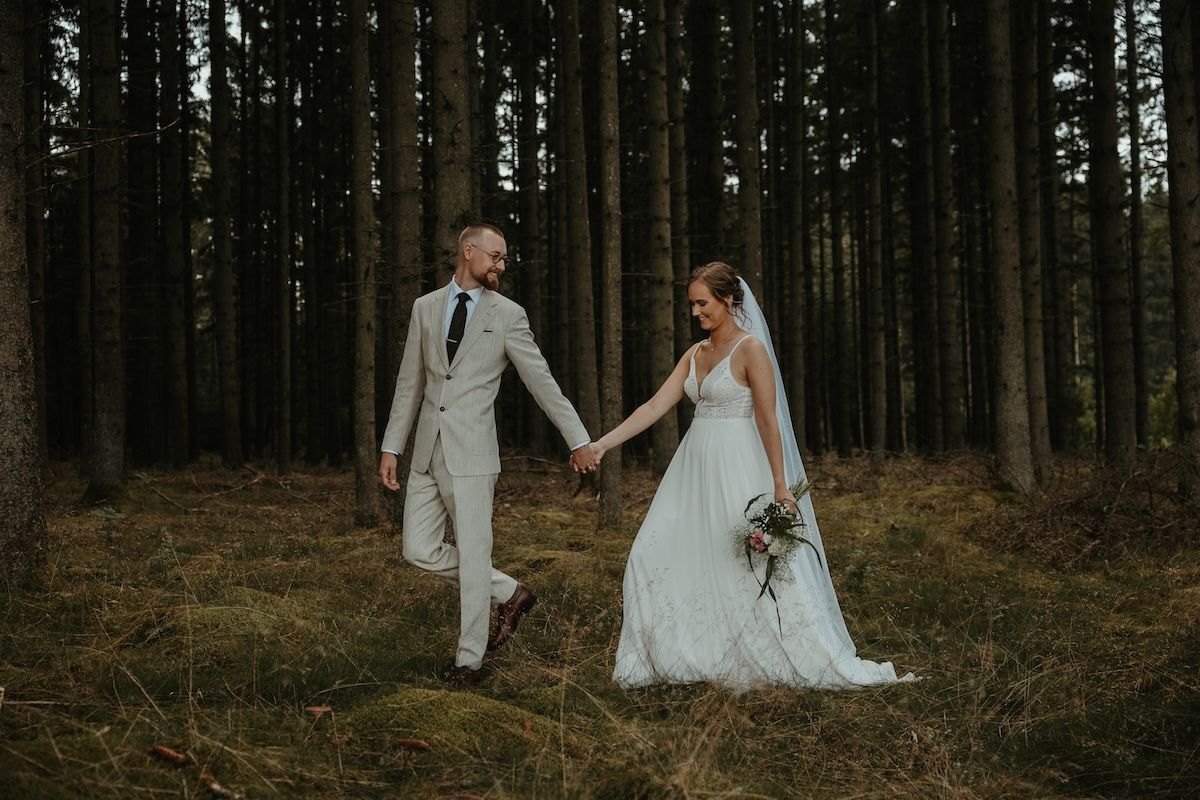 Brudpar går hand i hand genom en mossbeklädd skog. Fotograf Grevholm Photography.