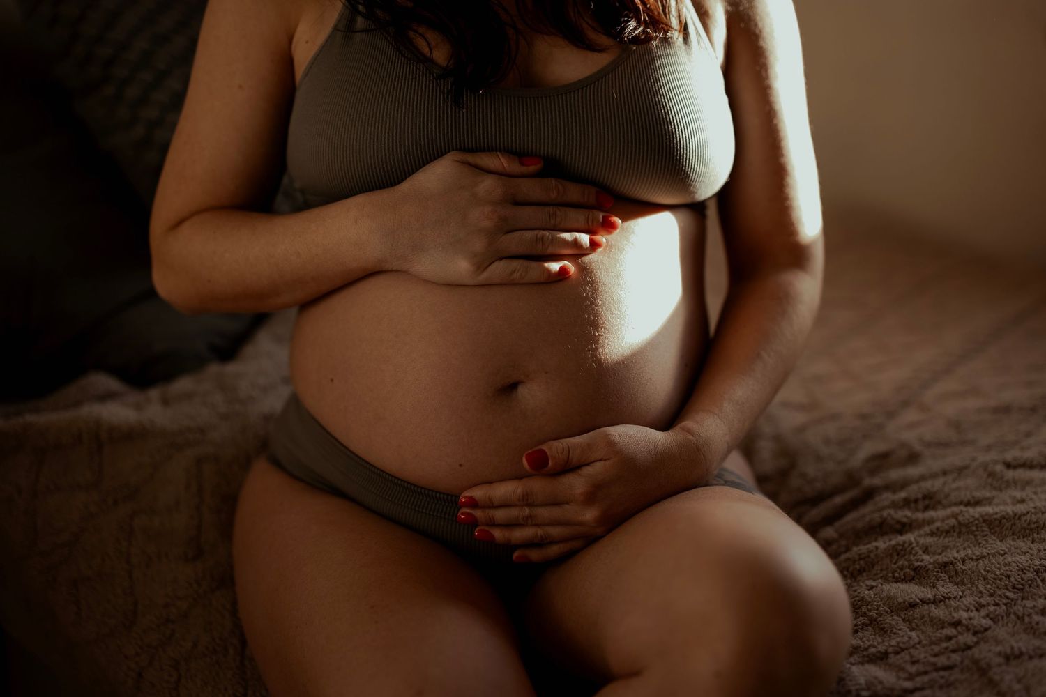 Gravid kvinna sitter på sängkanten iklädd underkläder under en gravidfotografering i hemmet. Foto: Grevholm Photography.