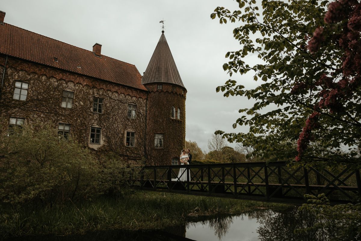 Brudpar står på gångbron framför Örtofta slott en mulen dag. Foto av Grevholm Photography.