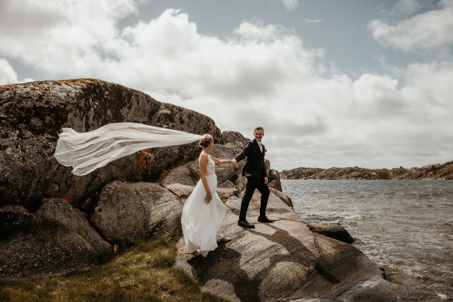Brudpar går hand-i-hand på klipporna vid havet i Orust och brudens slöja blåser högt i vinden.