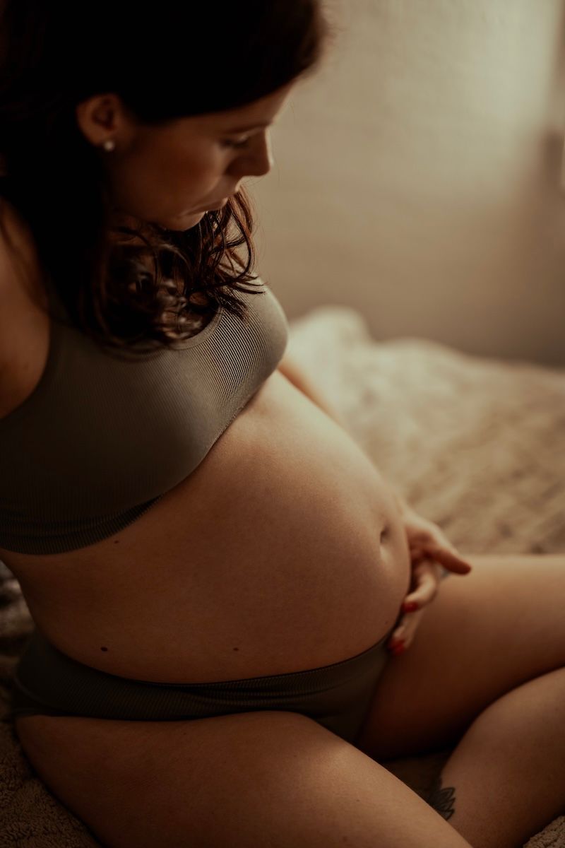 En gravid kvinna sitter på en sängkant och klappar ömt på sin bara mage. Foto av Grevholm Photography.
