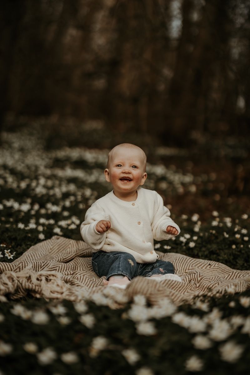 En glad bebis sitter på en filt bland vitsippor under en familjefotografering i naturen med Grevholm Photography.