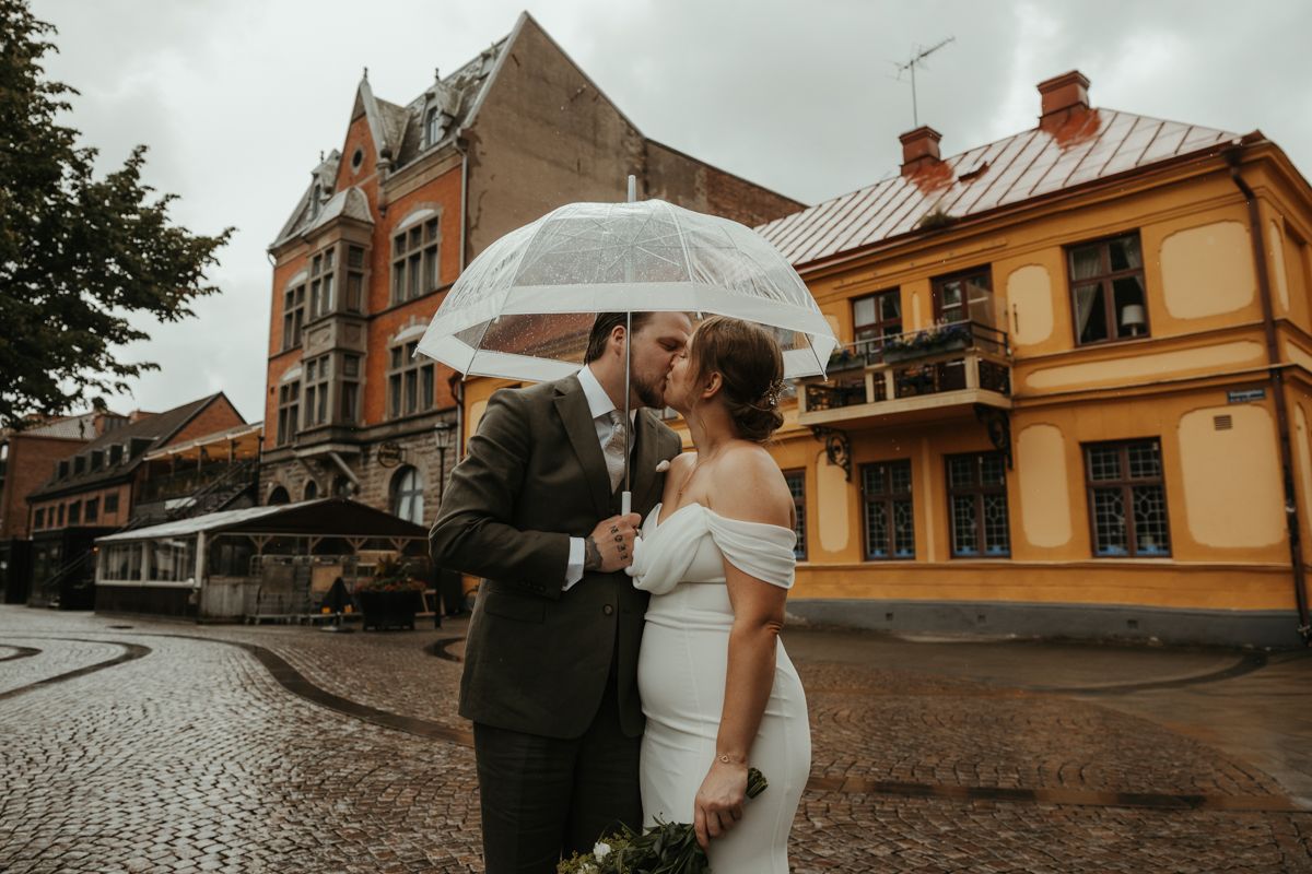 Brudpar står under ett paraply och pussas i ett regnigt Halmstad på deras bröllopsdag. Foto av Grevholm Photography.