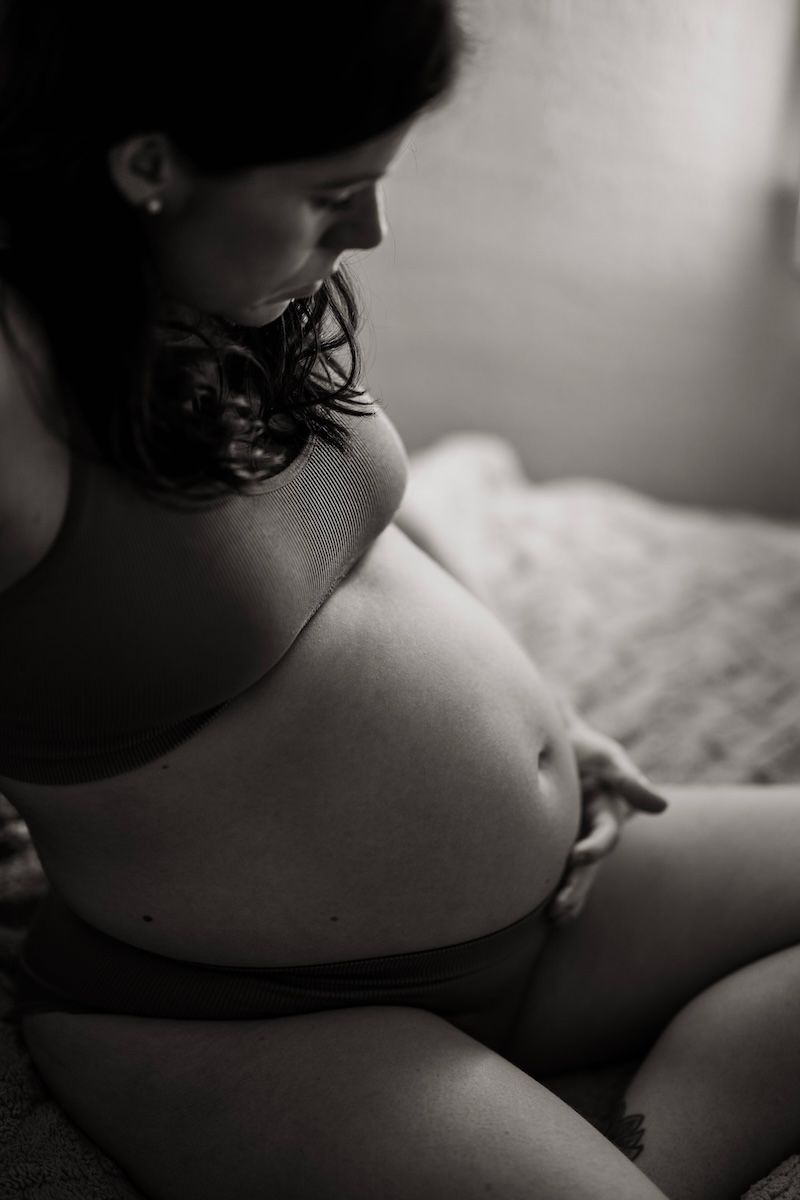 En gravid kvinna sitter på en sängkant och klappar på sin mage. Foto: Grevholm Photography.