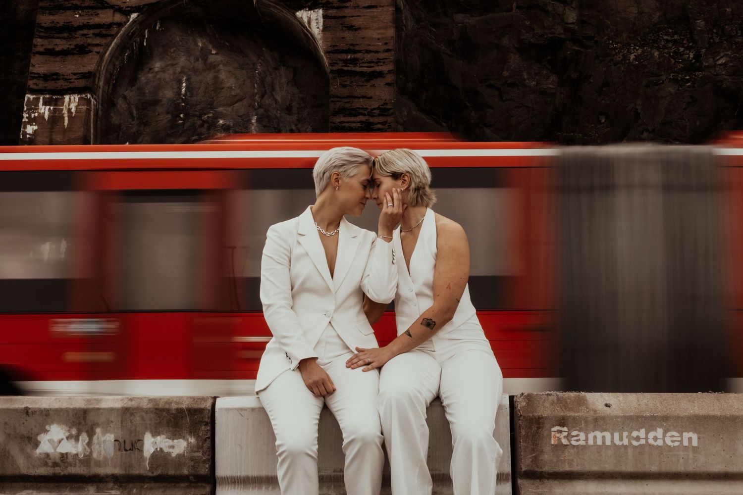 Brudpar sitter på en betongmur när en en röd buss kör förbi bakom dem i Stockholm. Foto av Grevholm Photography.