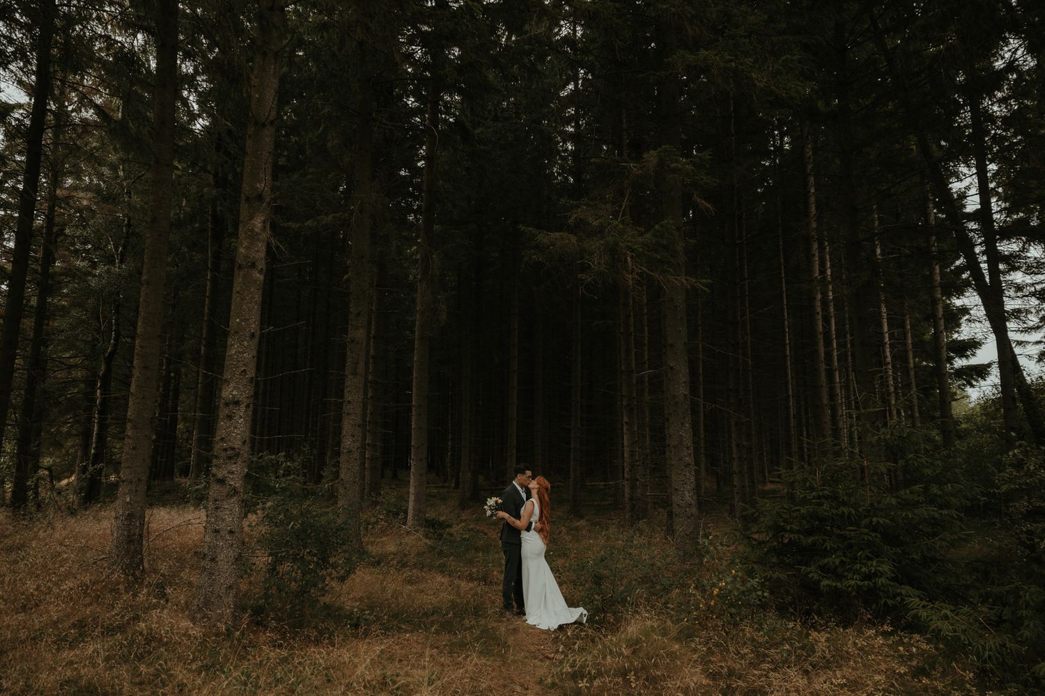 Brudpar står i en barrskog och pussas. Foto av Grevholm Photography.