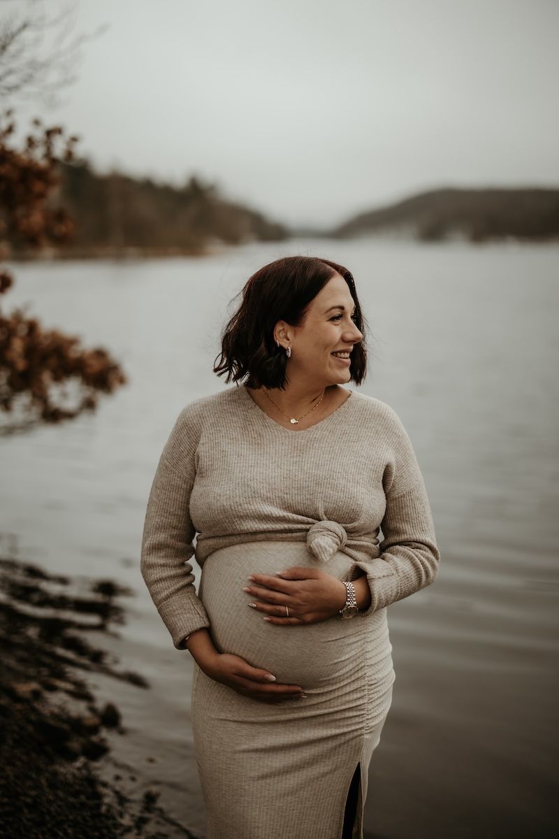 En gravid kvinna står vid vattenbrynet av en sjö en disig dag i januari och håller om sin mage. Foto av Grevholm Photography