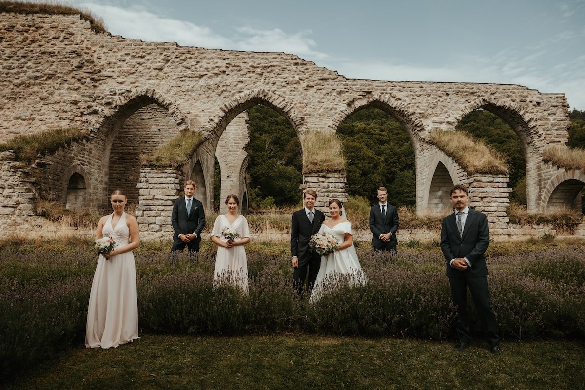 Brudpar och deras brudfölje poserar kaxigt framför Alvastra klosterruin i Ödeshög. Fotograf Grevholm Photography.