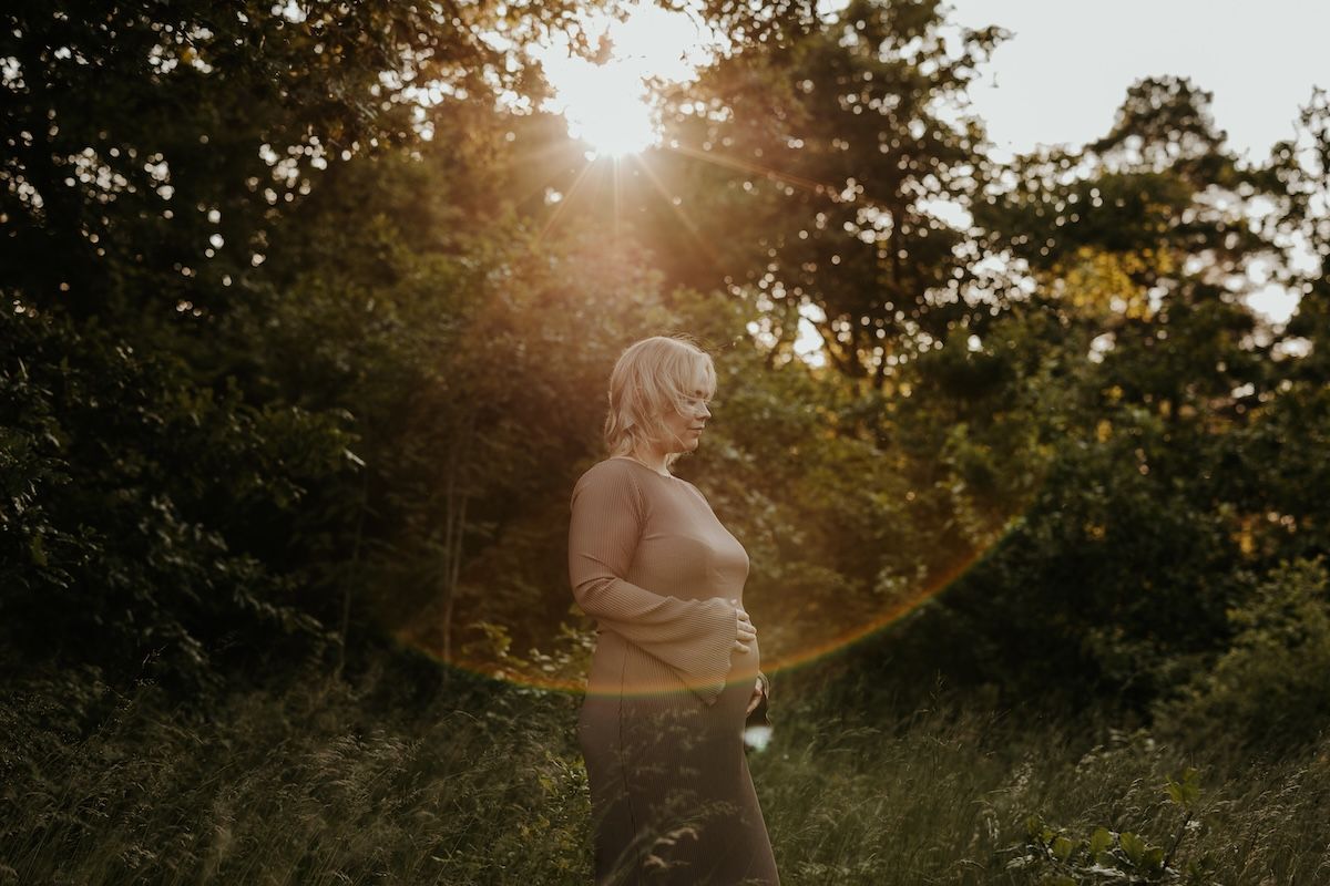 En gravid kvinna iklädd en klänning står på ett fält med högt gräs och håller om sin mage under solnedgång. Foto av Grevholm Photography