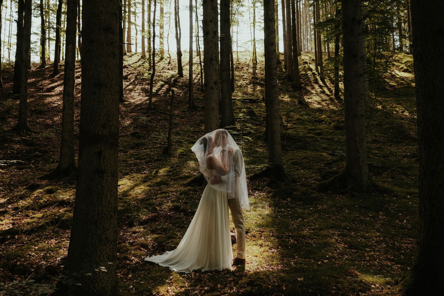 Brudpar kysser varandra under brudslöjan ståendes i skogen med solen bakom dem. Foto: Grevholm Photography.