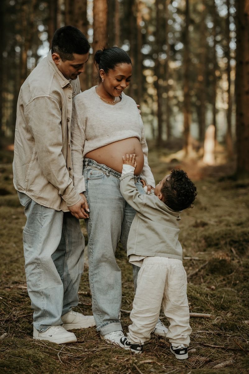 Expecting parents and young child share tender moment touching pregnant belly in wooded forest setting.
