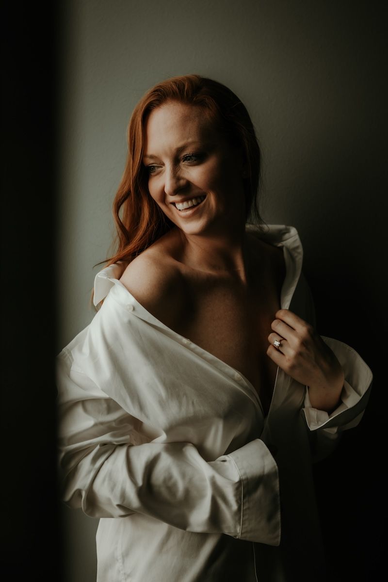 Kvinna iklädd en vit skjorta blickar ut genom ett fönster och ser glad ut under en boudoirfotografering i Skåne. Foto: Grevholm Photograpy.