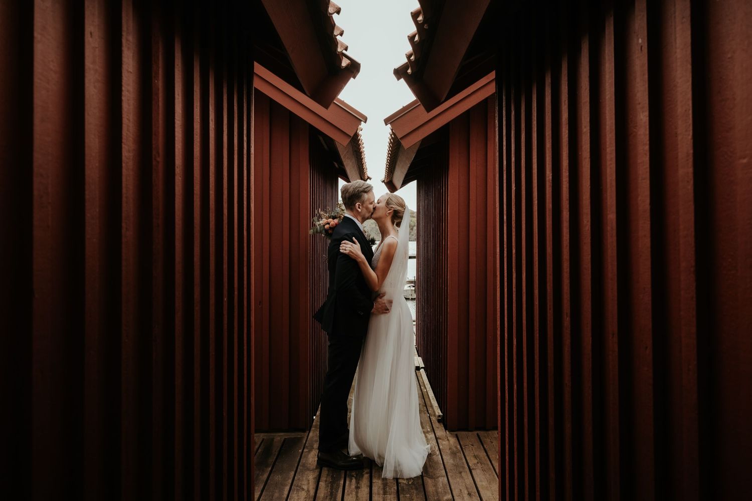 Brudpar står inne bland röda båthus och kysser varandra under deras bröllop på västkusten. Foto av Grevholm Photography.