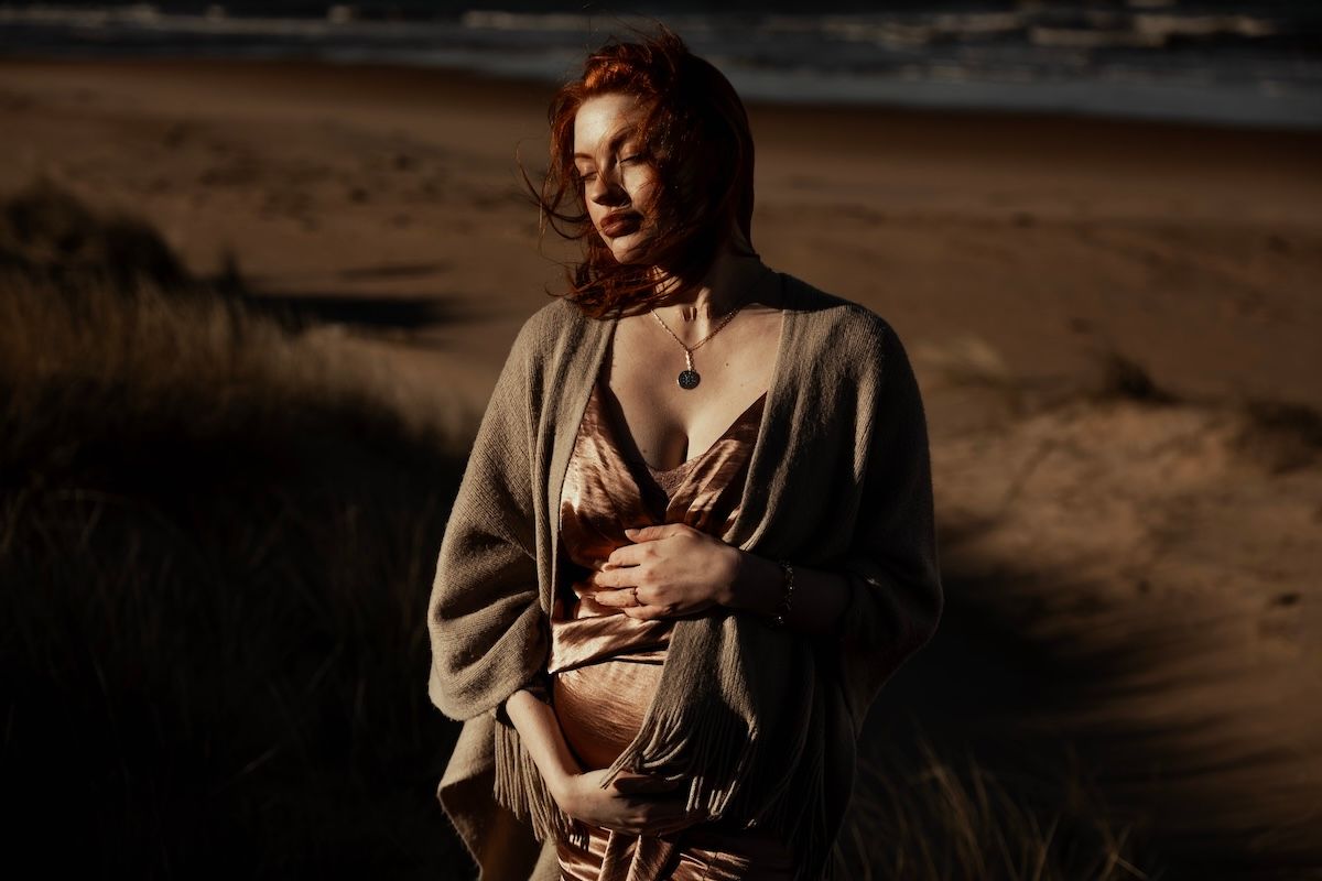 En gravid kvinna iklädd en rosa klänning står på stranden och klappar sin mage under en gravidfotografering i Laholm. Grevholm Photography