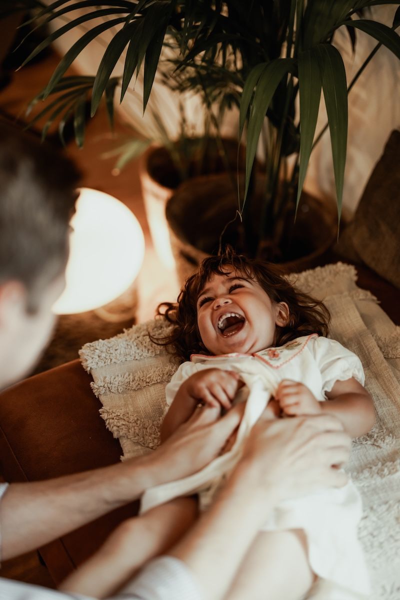 En pappa kittlar sin dotter ligger ner på en soffa och skrattar under en lifestylefotografering i Skåne med Grevholm Photography